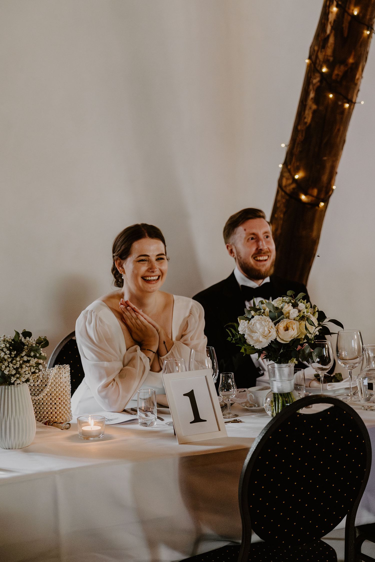 Wedding reception setting with candlelit table and rustic wooden beams decorated with floral arrangements and place settings.