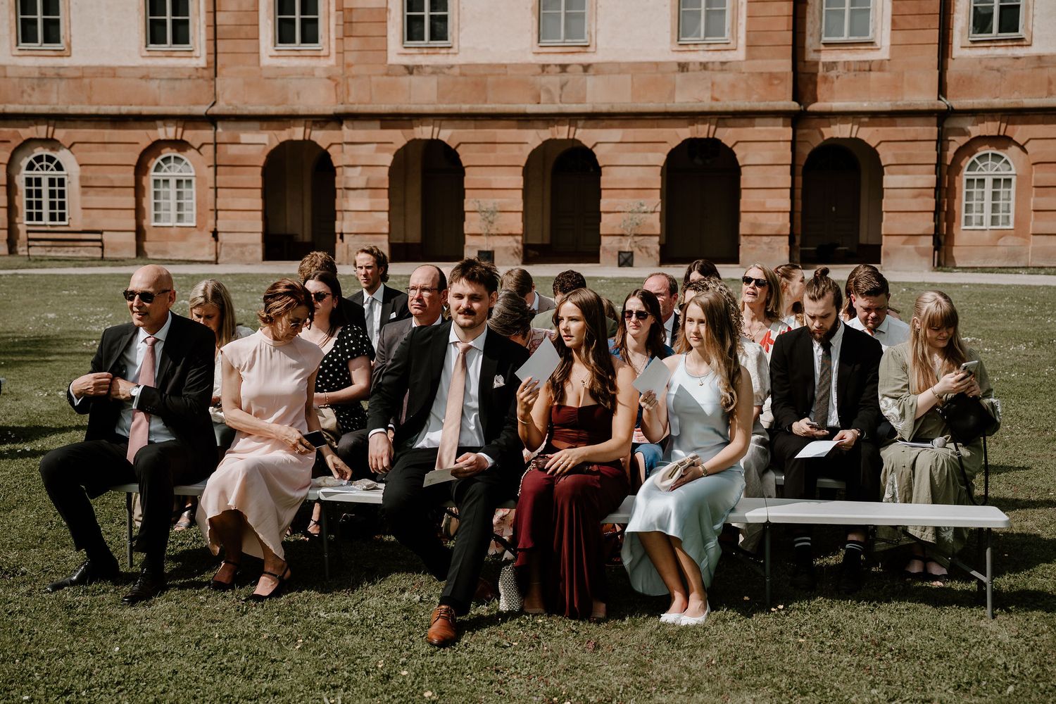 Wedding party sits casually on benches outdoors against historic brick building facade on sunny afternoon.