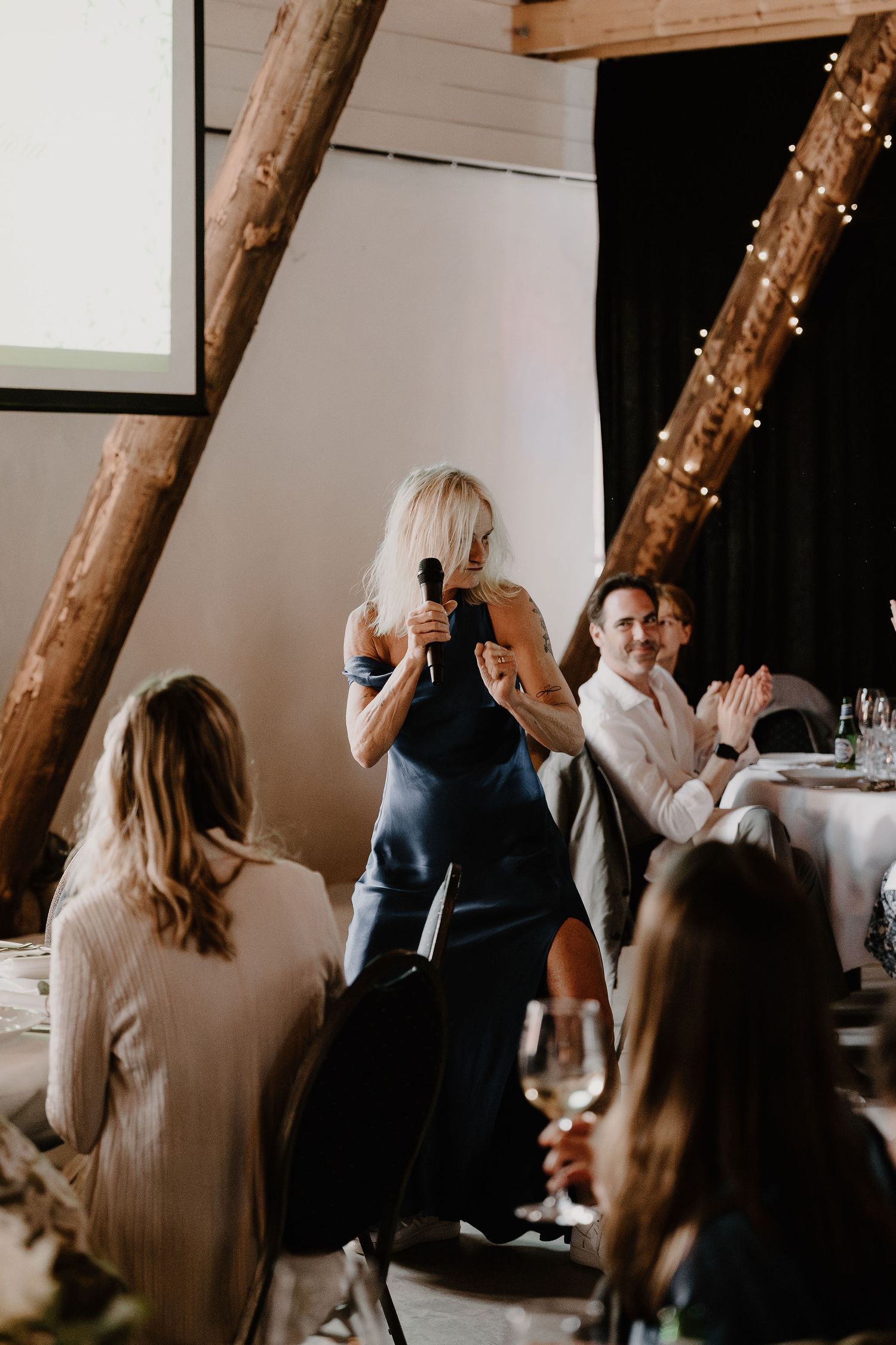 People gather for a celebration dinner in a rustic venue with wooden beams and string lights above.