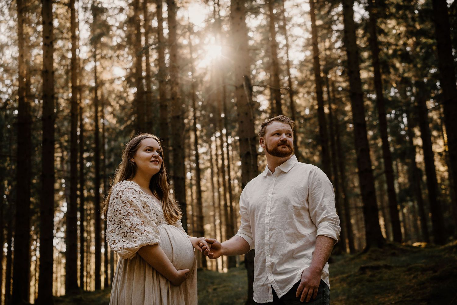 A romantic couple holds hands in a sunlit forest clearing during golden hour, with tall evergreen trees surrounding them.