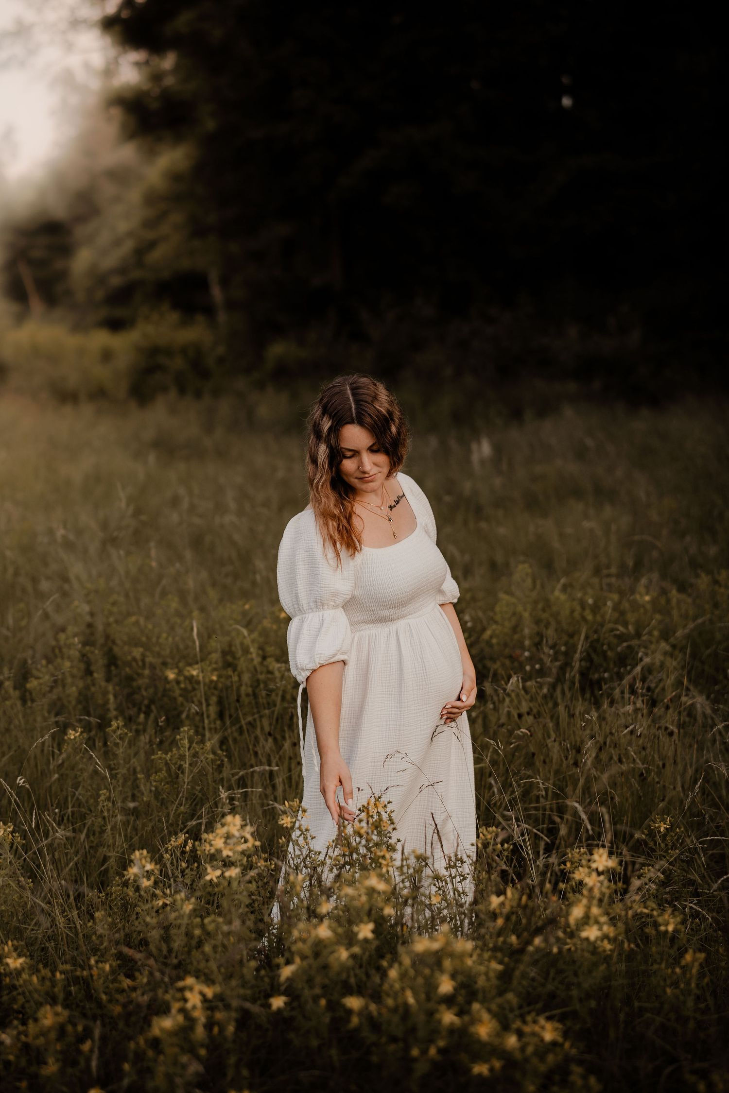 A woman in a flowing white dress stands in a golden meadow at sunset while cradling her pregnant belly.