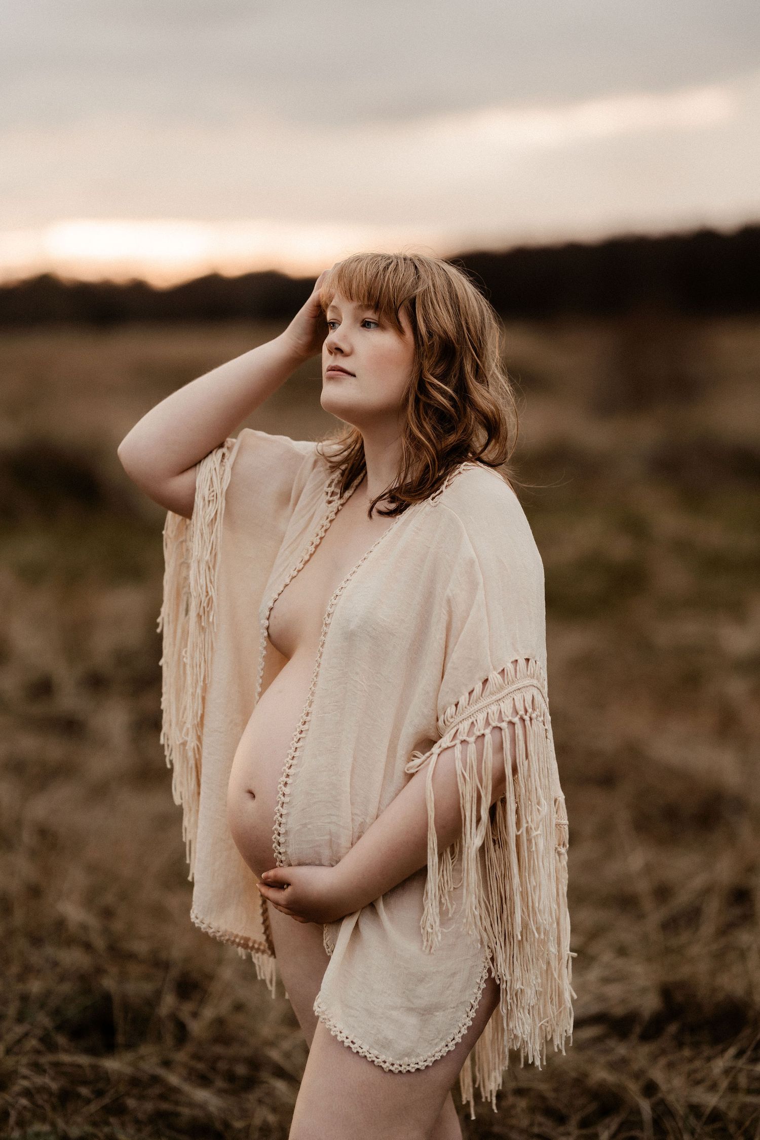 A pregnant woman poses in a cream fringed shawl against a moody sunset backdrop in a rustic outdoor setting.