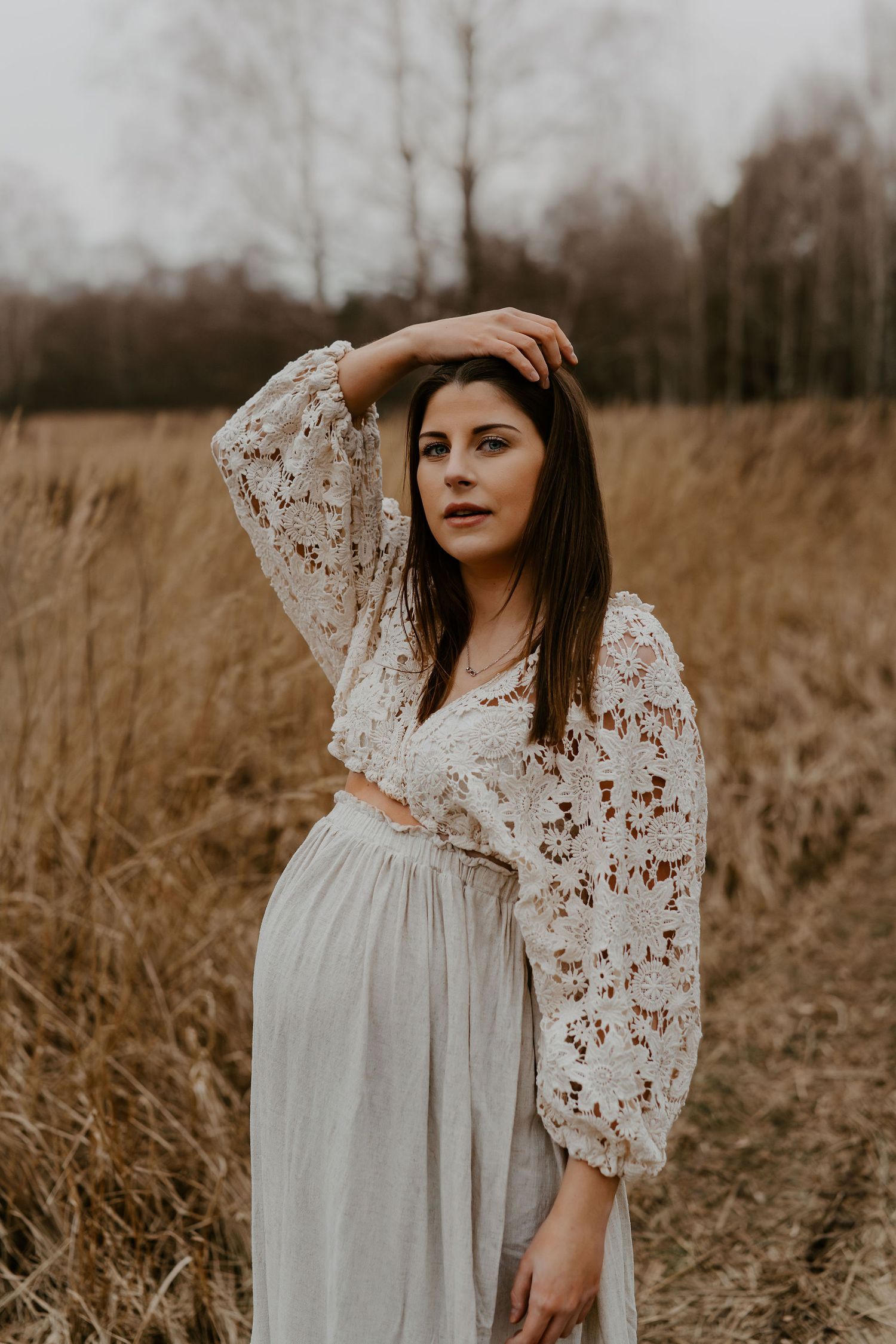 A dreamy maternity photo of someone in a white lace dress poses thoughtfully in a golden autumn field.