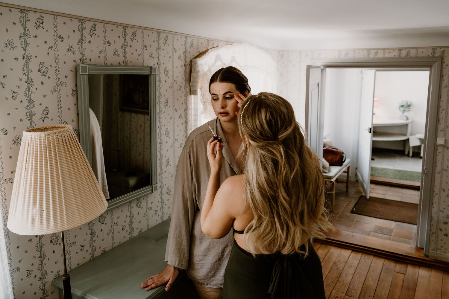 A couple shares an intimate moment in front of a mirror in a bedroom with floral wallpaper and a lamp.