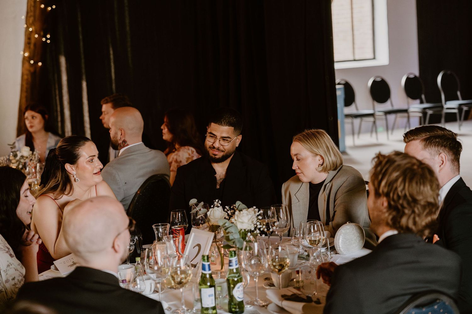 Wedding reception guests laugh and socialize while seated at a dimly lit table decorated with wine glasses and centerpieces.