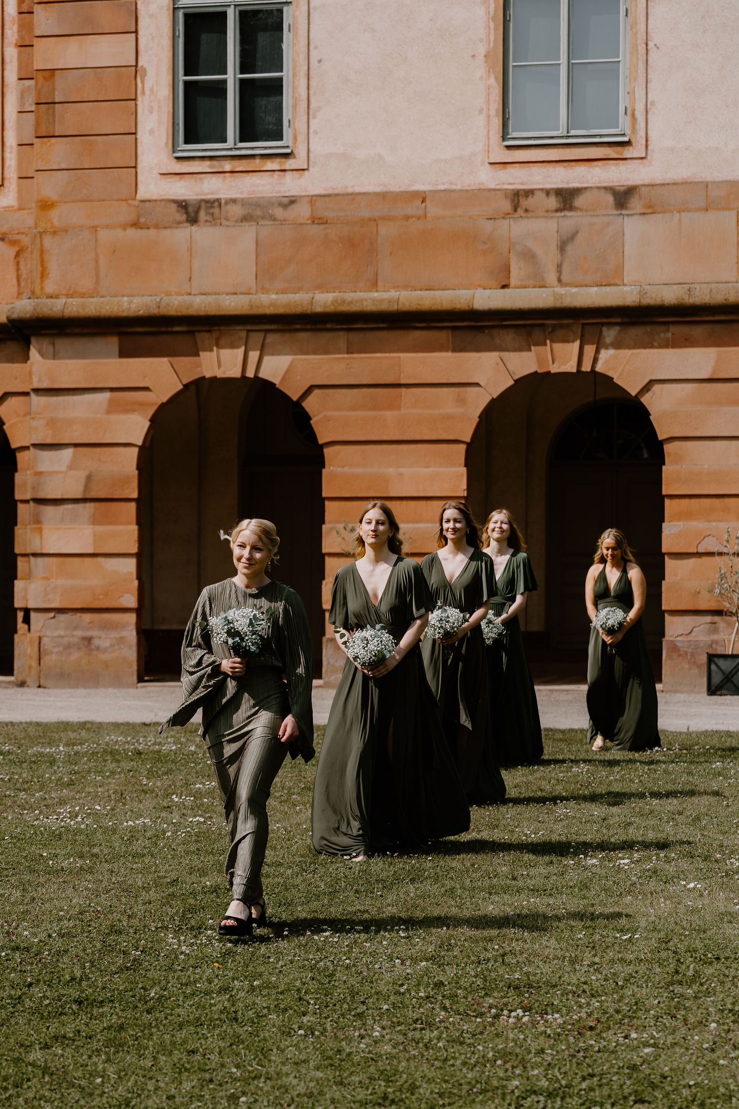 Group of people in dark clothing gather on a grassy lawn outside a historic building with multiple windows.