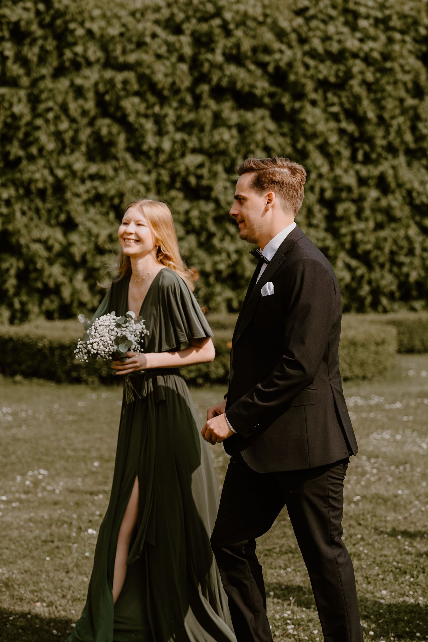 A couple walking together on a lawn, wearing formal attire, with the woman in a green dress holding baby's breath flowers.