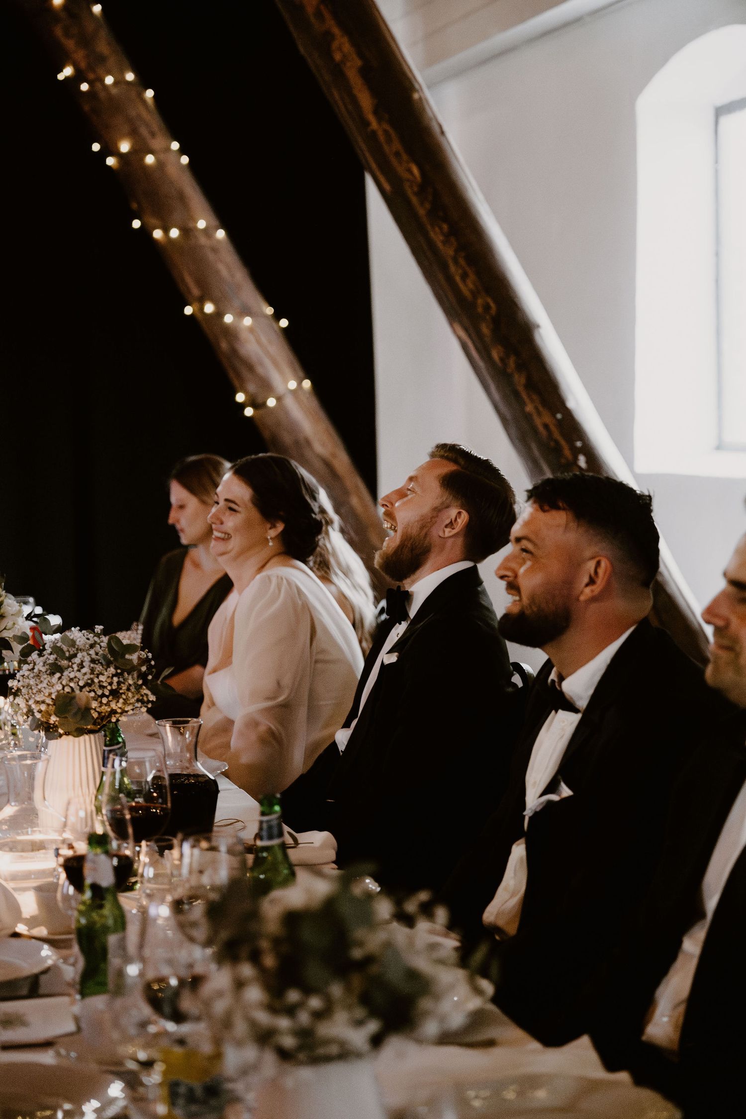 Guests seated at an elegant wedding reception table decorated with string lights and floral centerpieces.