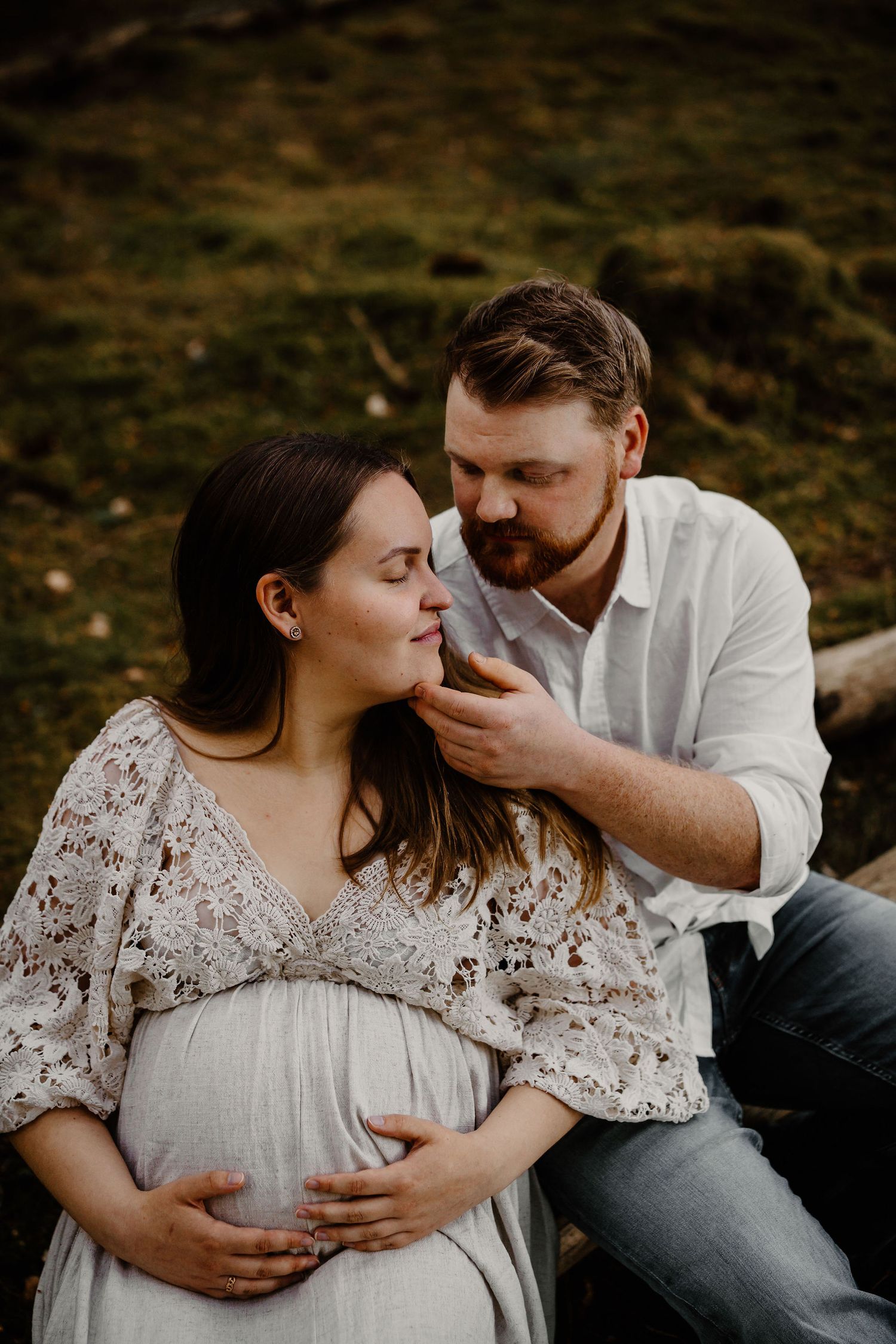 Intimate maternity photo shows couple sitting outdoors with pregnant woman wearing flowy white dress and man in casual white shirt.