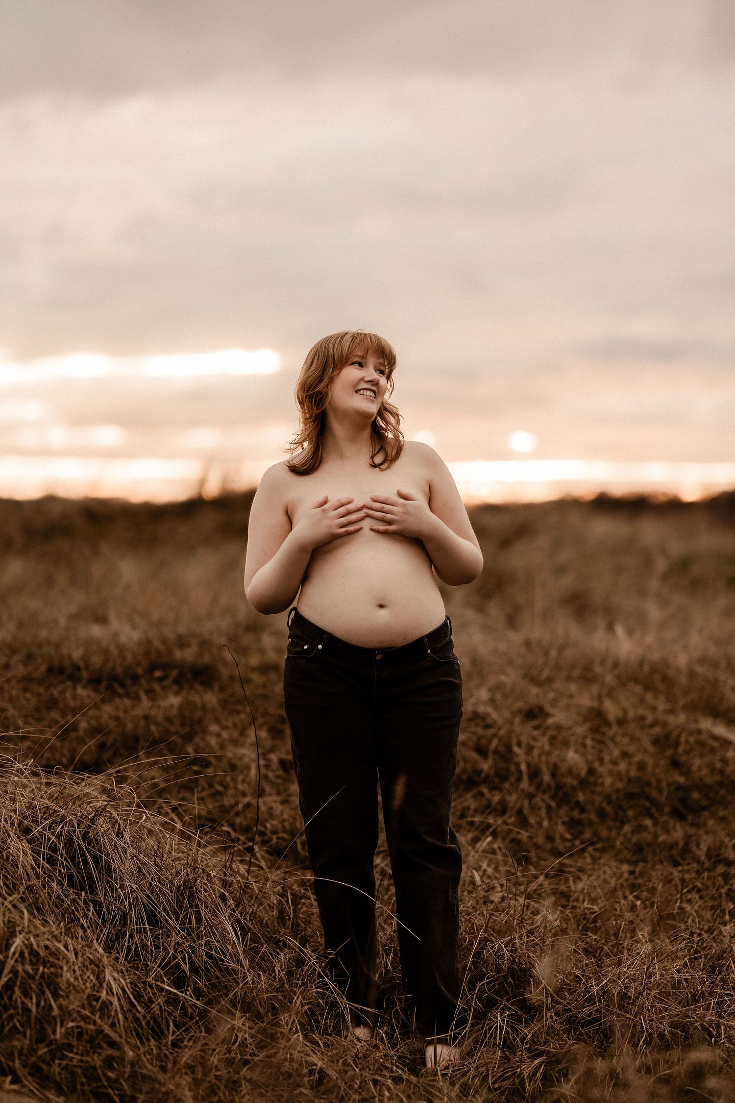 A silhouetted figure stands in a golden field at sunset in a dreamy sepia-toned maternity portrait.