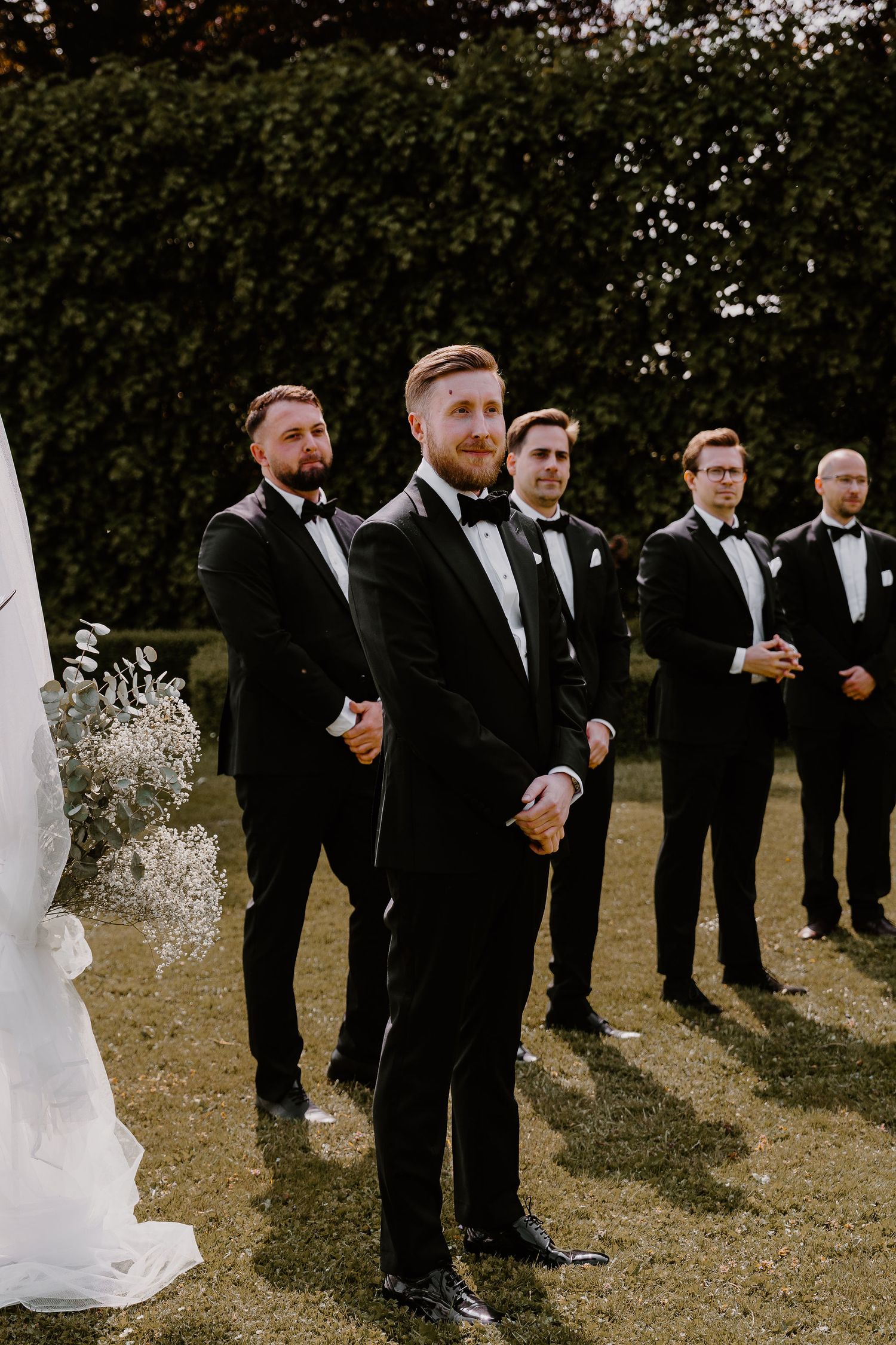 Groomsmen in classic black tuxedos stand in line during outdoor wedding ceremony with green hedges in background.