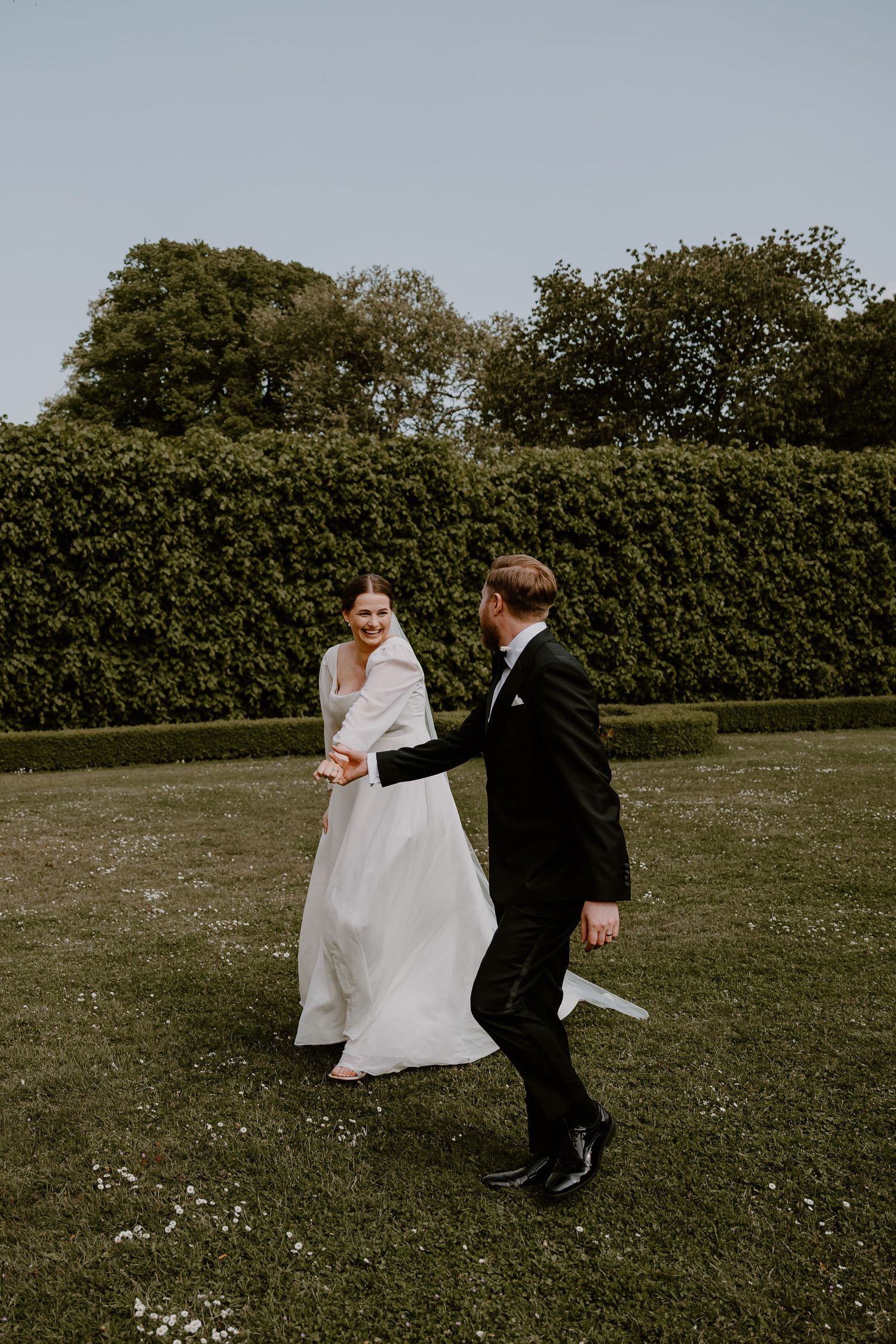A couple dances together on a lawn with manicured hedges and trees in the background during their wedding celebration.