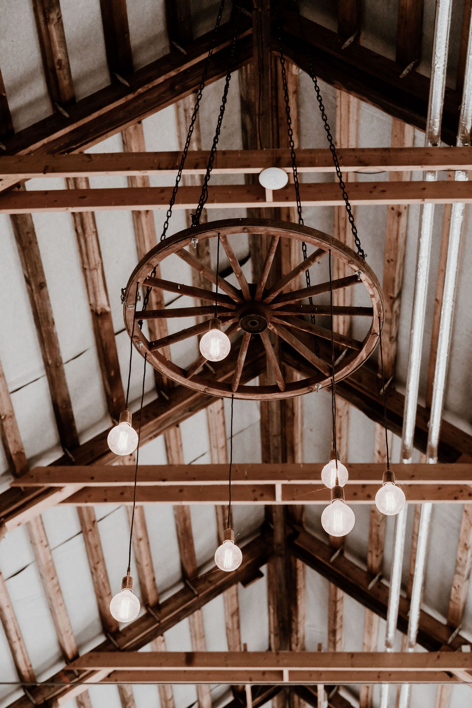 Rustic wagon wheel chandelier with hanging light bulbs illuminates wooden beamed ceiling in sepia tones.