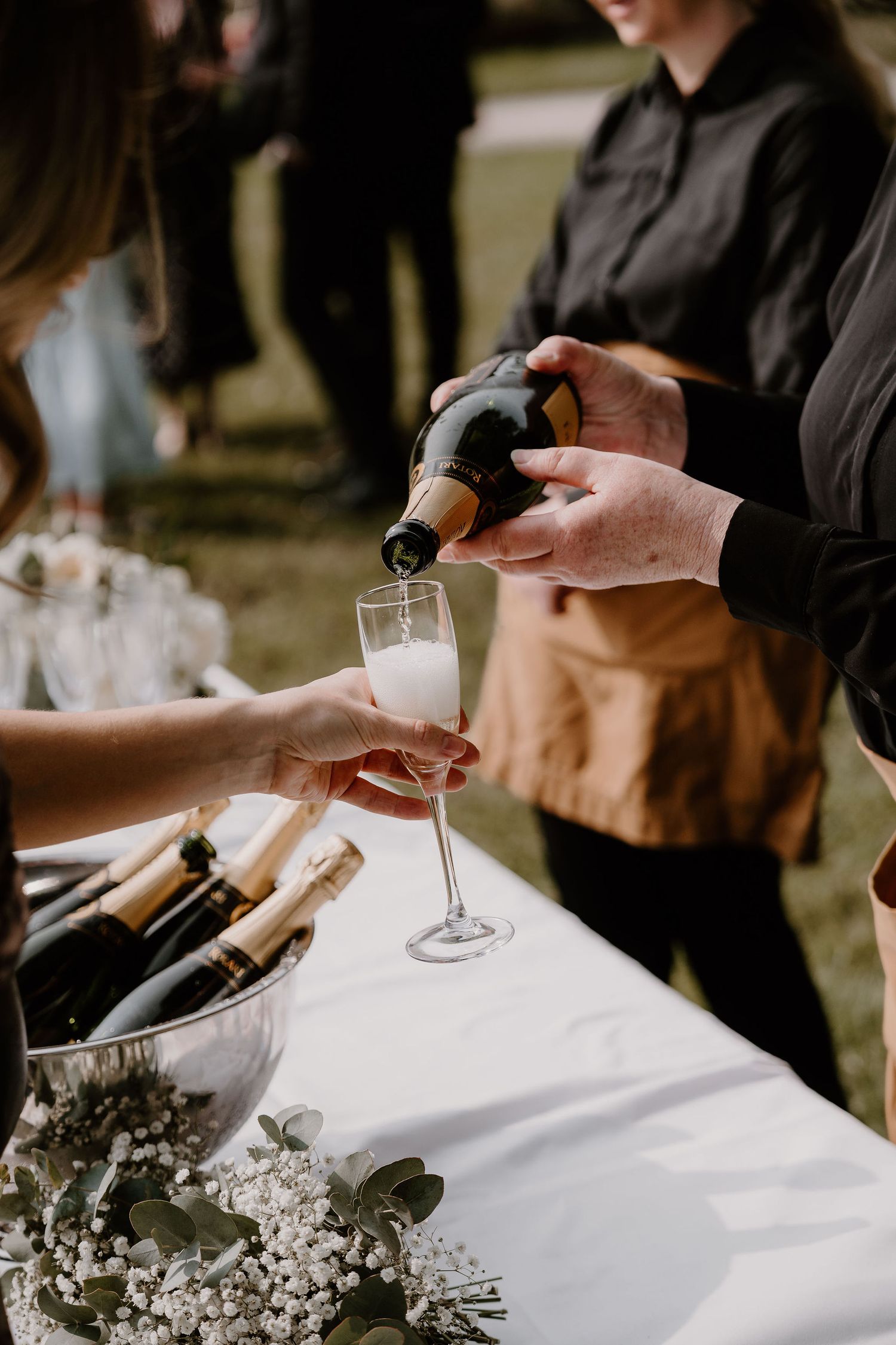 Champagne being poured into a glass at an outdoor event with white tablecloths and baby's breath floral decorations.