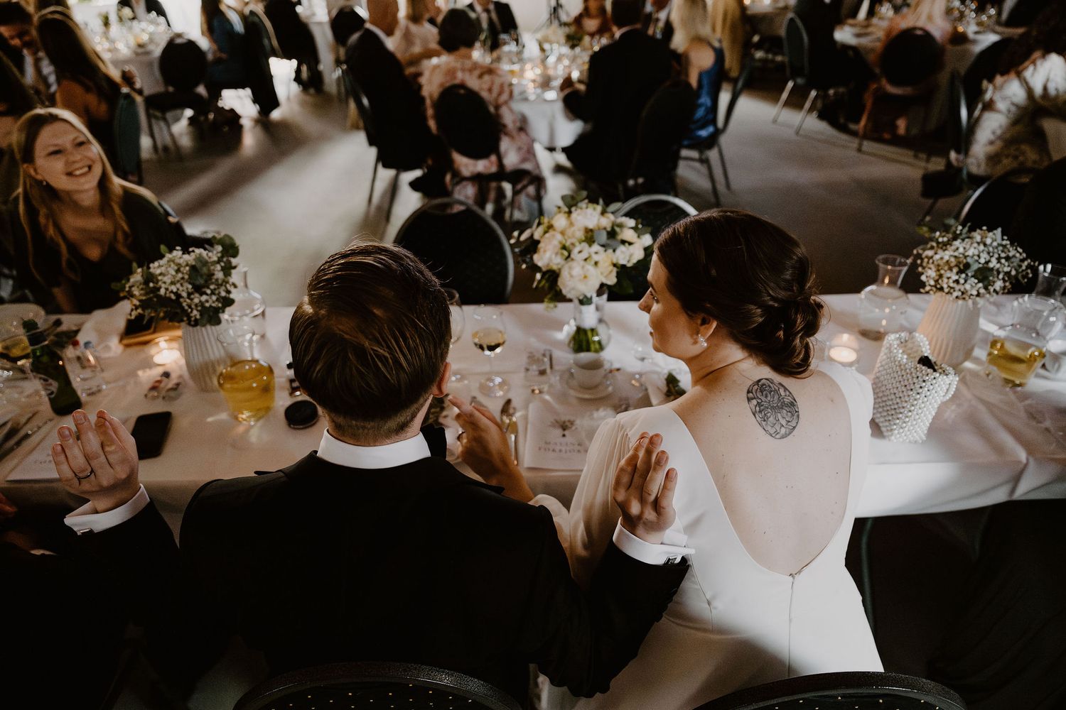 Couple sits at elegant wedding reception table with white decor and floral arrangements while guests dine in background.