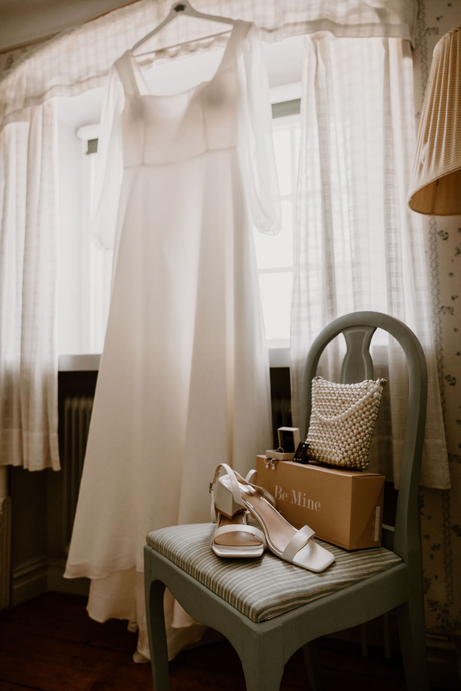 White wedding dress hanging near window with bridal heels and decorative pillow on gray chair in soft natural light.