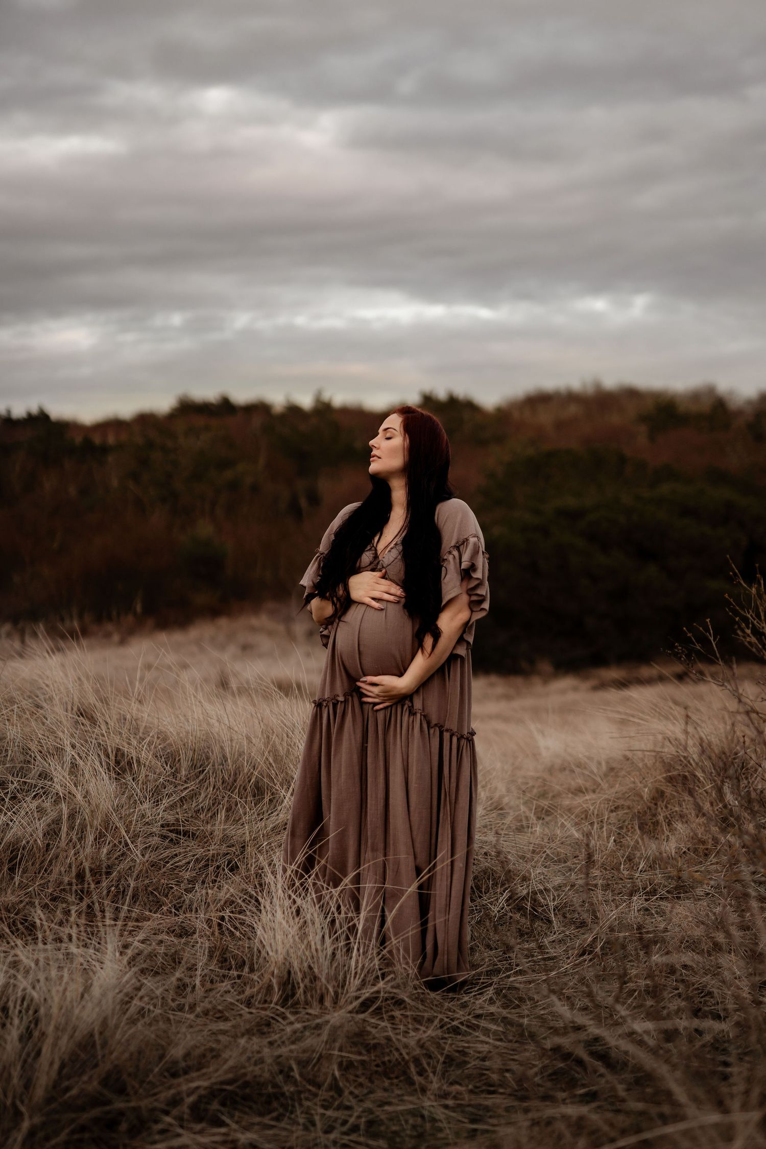 A pregnant woman in a flowing brown dress stands in a serene field at sunset with dramatic cloudy skies overhead.