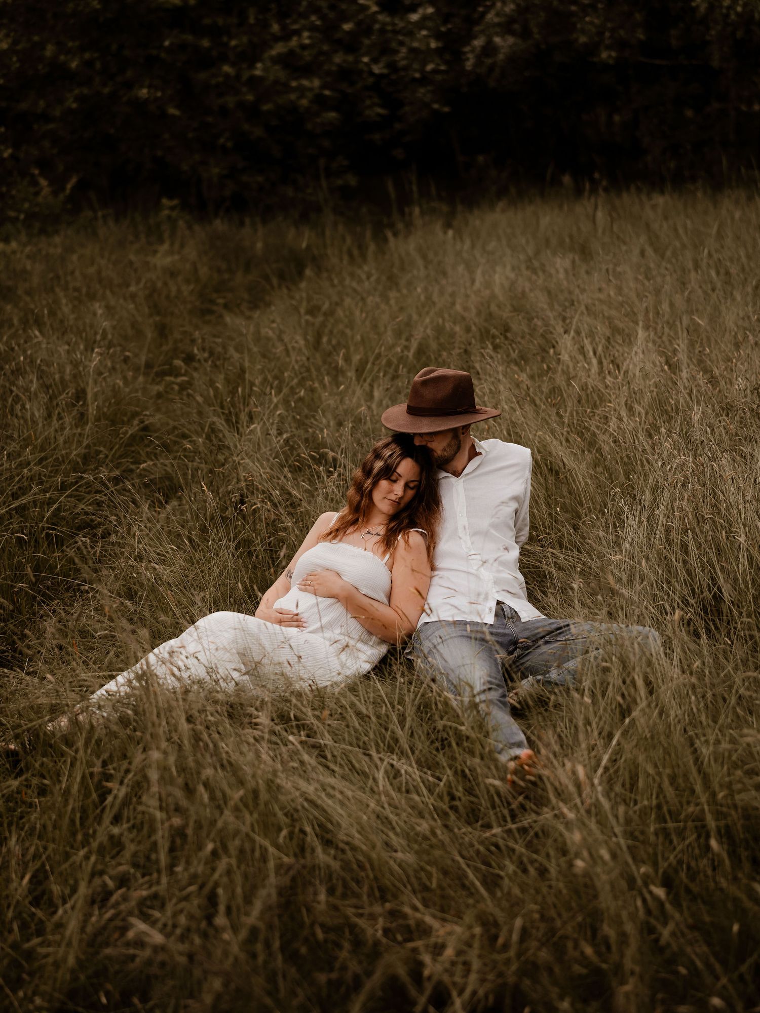 A couple sits intimately together in a field of tall grass during a romantic outdoor photoshoot at sunset.