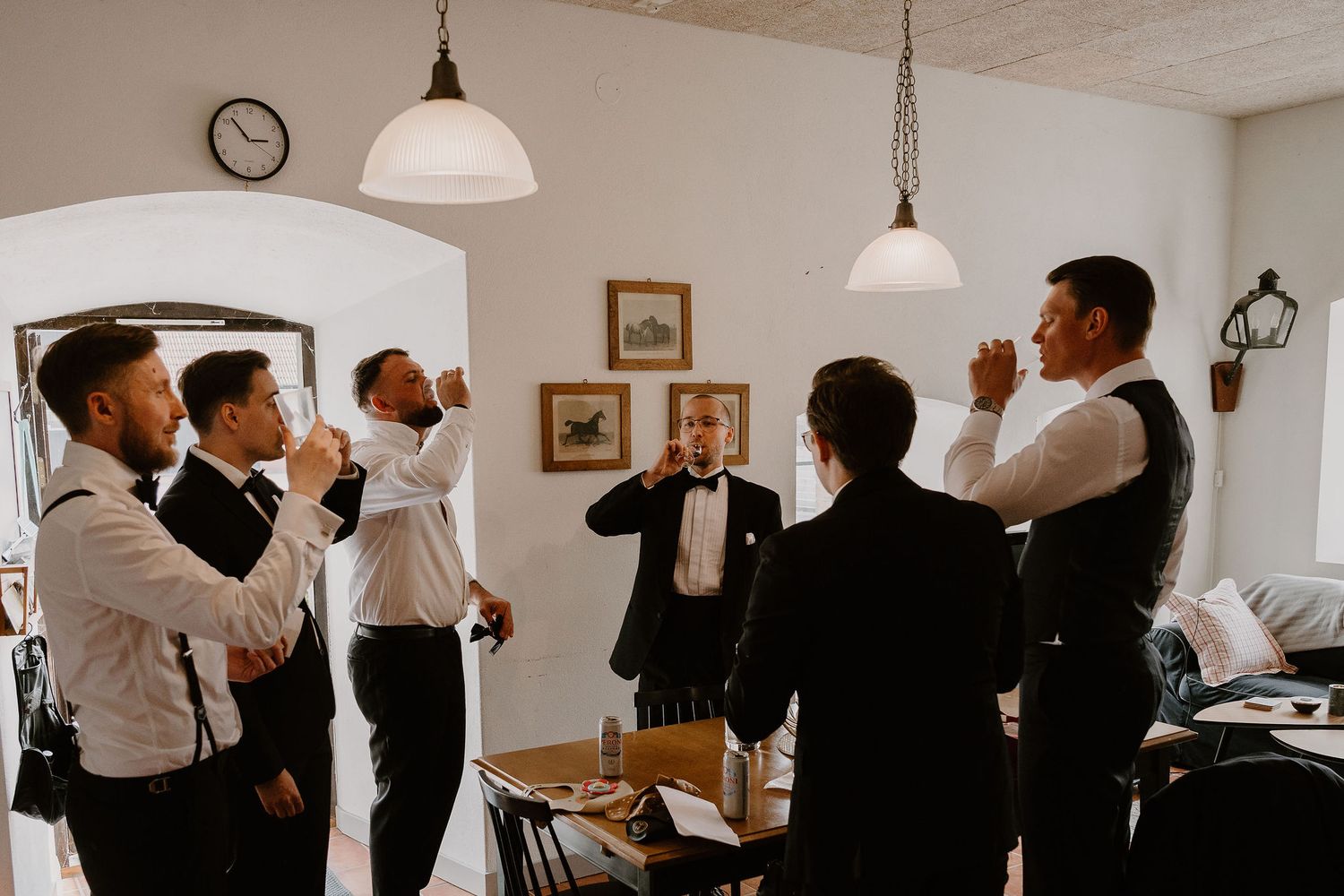 Wedding party members in formal attire chat and mingle at an indoor reception venue with classic pendant lighting.