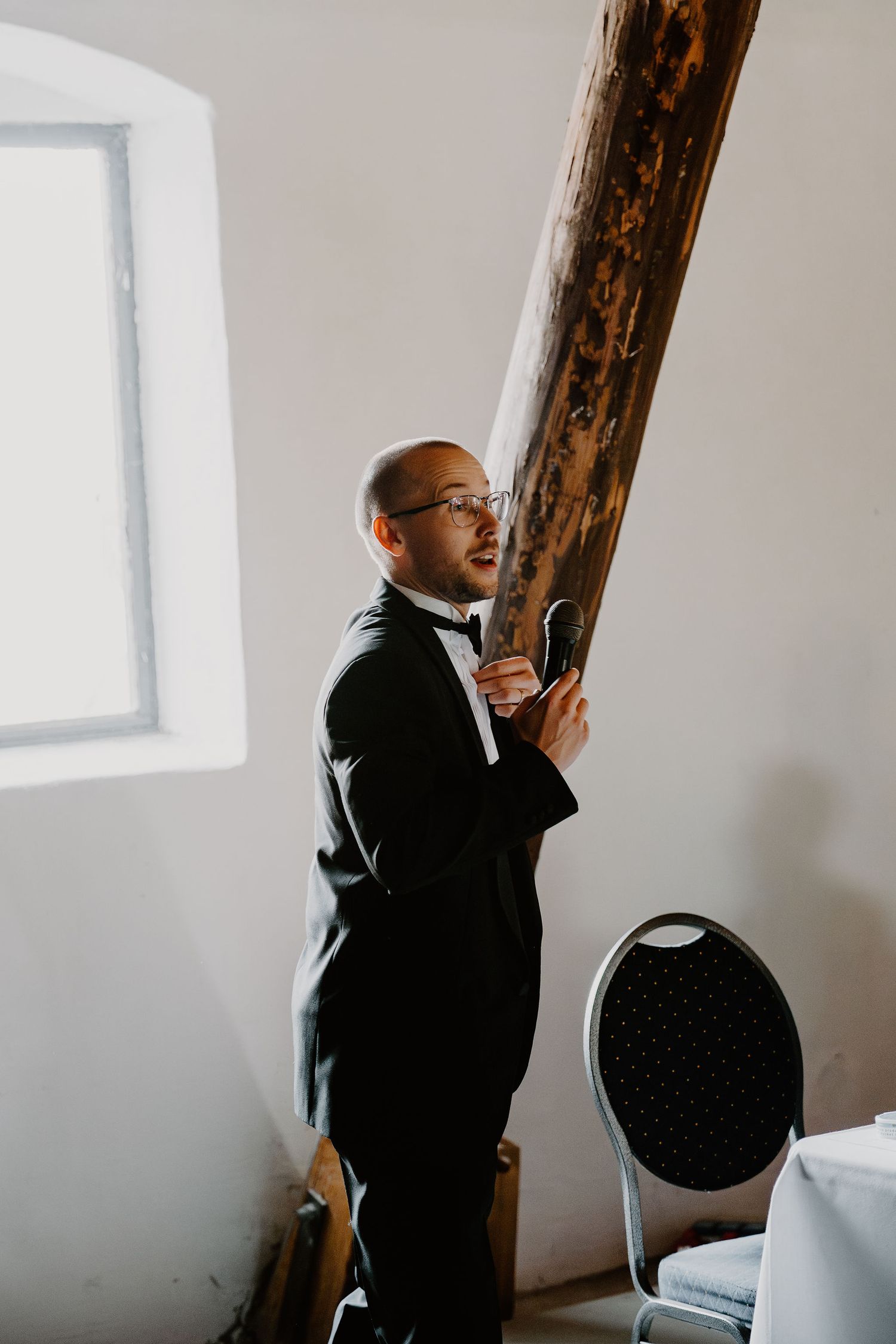 Professional in a black suit stands contemplatively by a wooden beam in a minimalist white room.