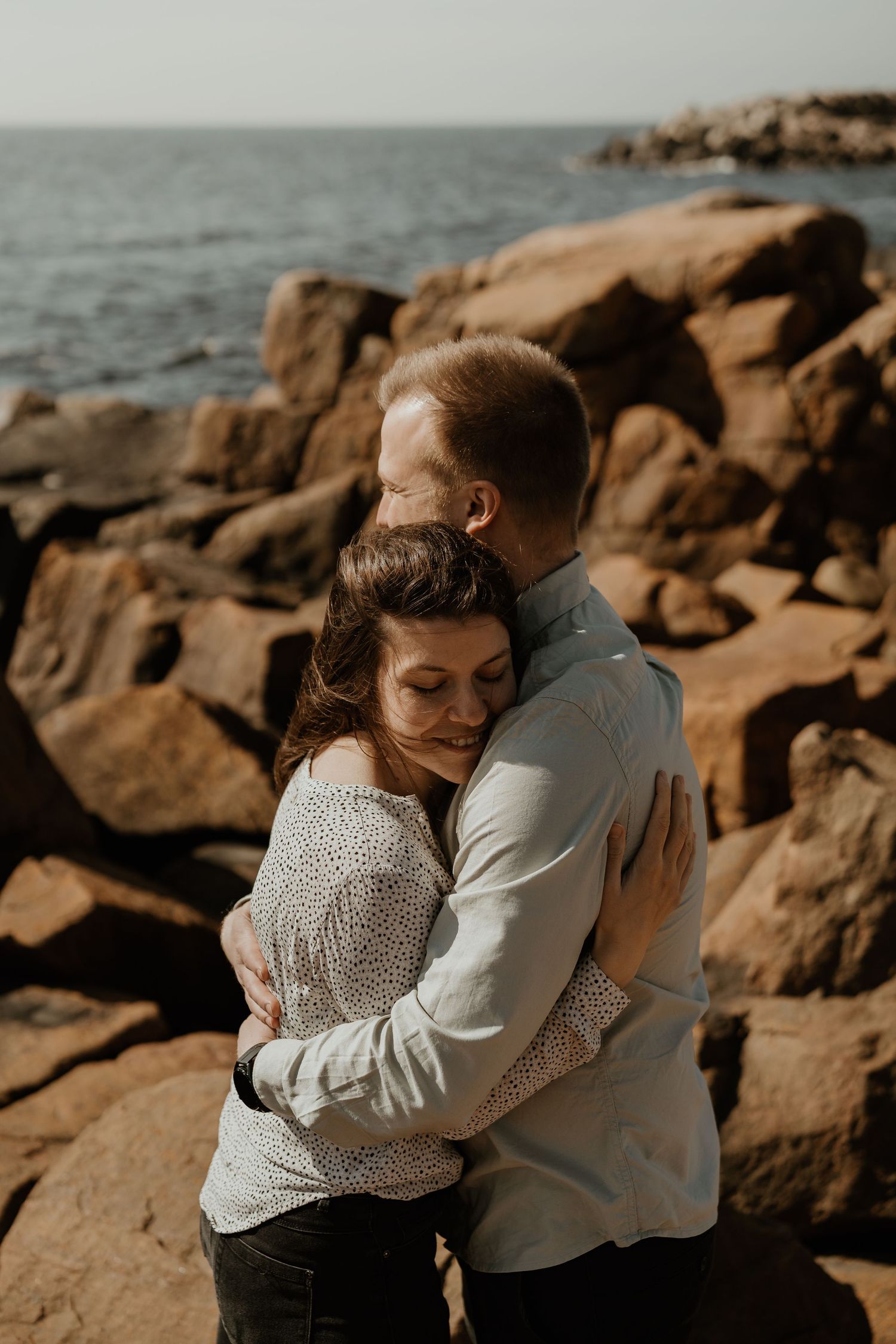 A couple shares an intimate embrace on rocky coastline during golden hour as waves crash against the shore.
