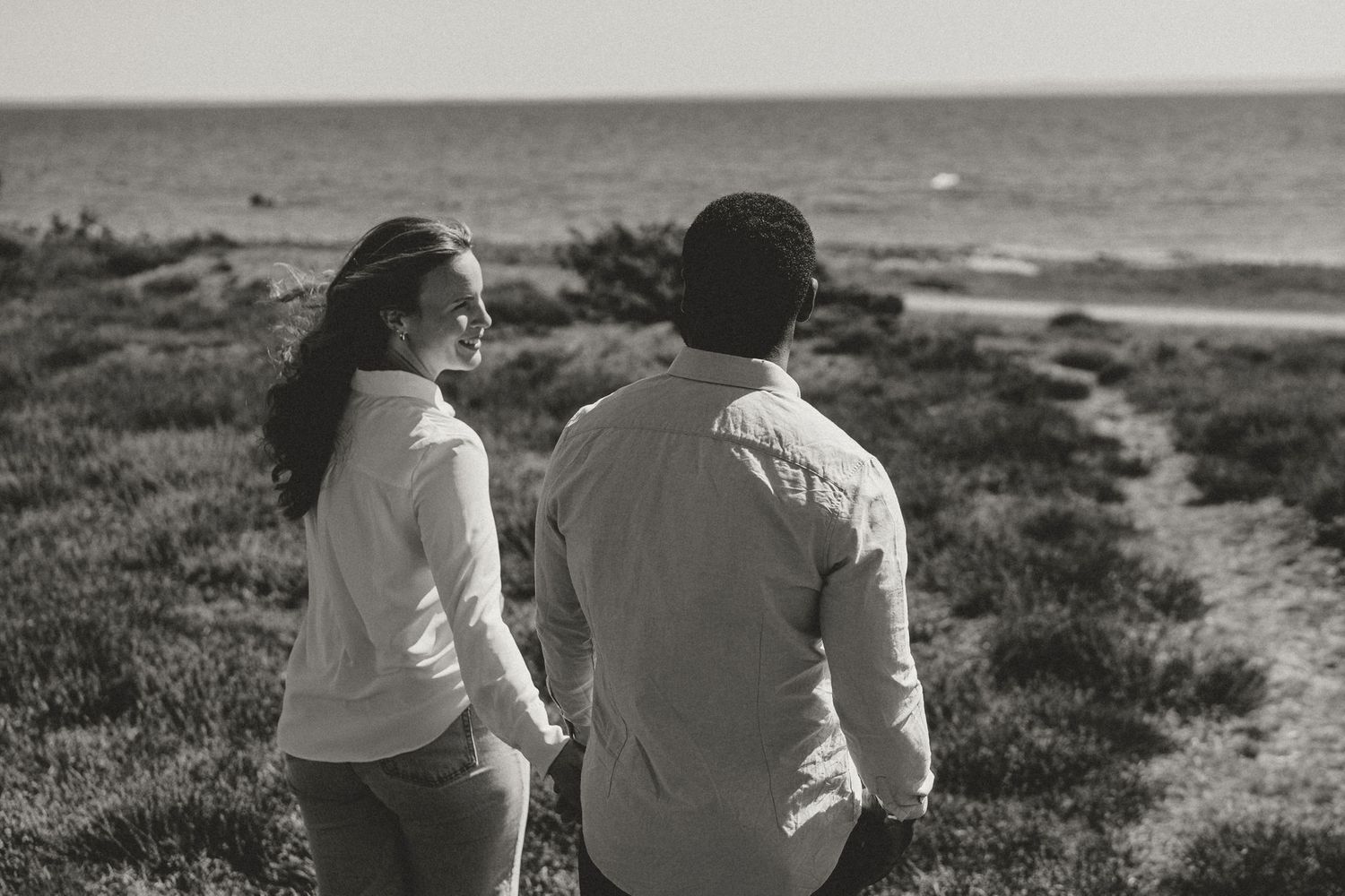 A couple sits together on grassy seaside cliffs overlooking the ocean in black and white.