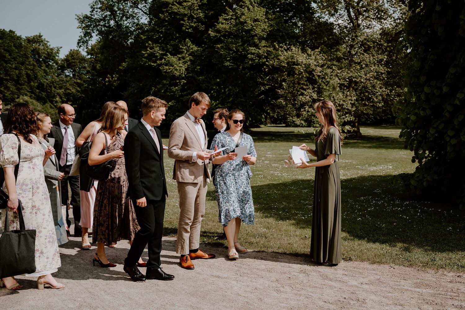 A small group gathers outdoors for a summer wedding ceremony in a sunlit park setting with lush trees in the background.