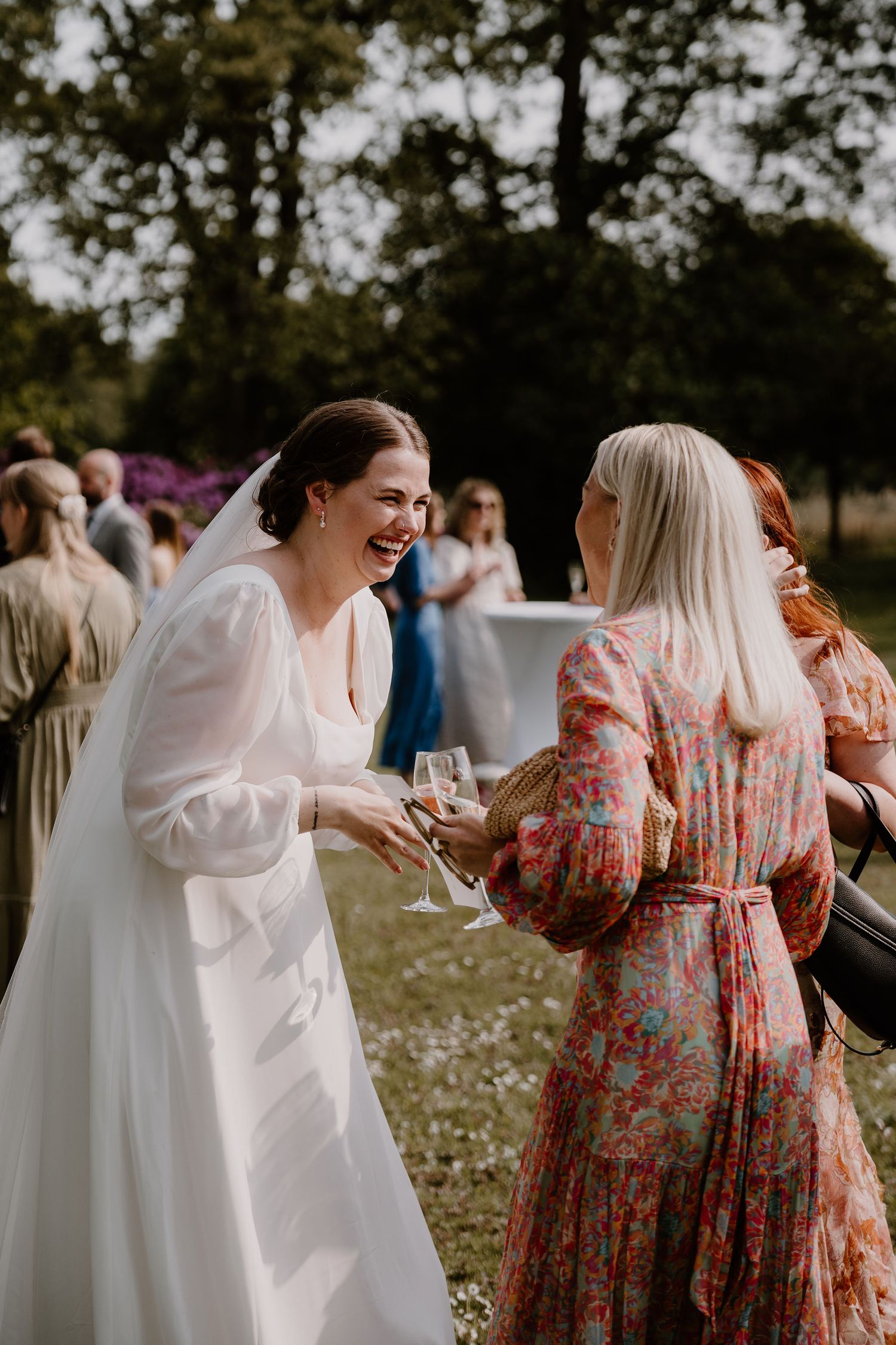 Two guests share a joyful moment at an outdoor wedding reception, with one wearing a white gown and another in floral print.
