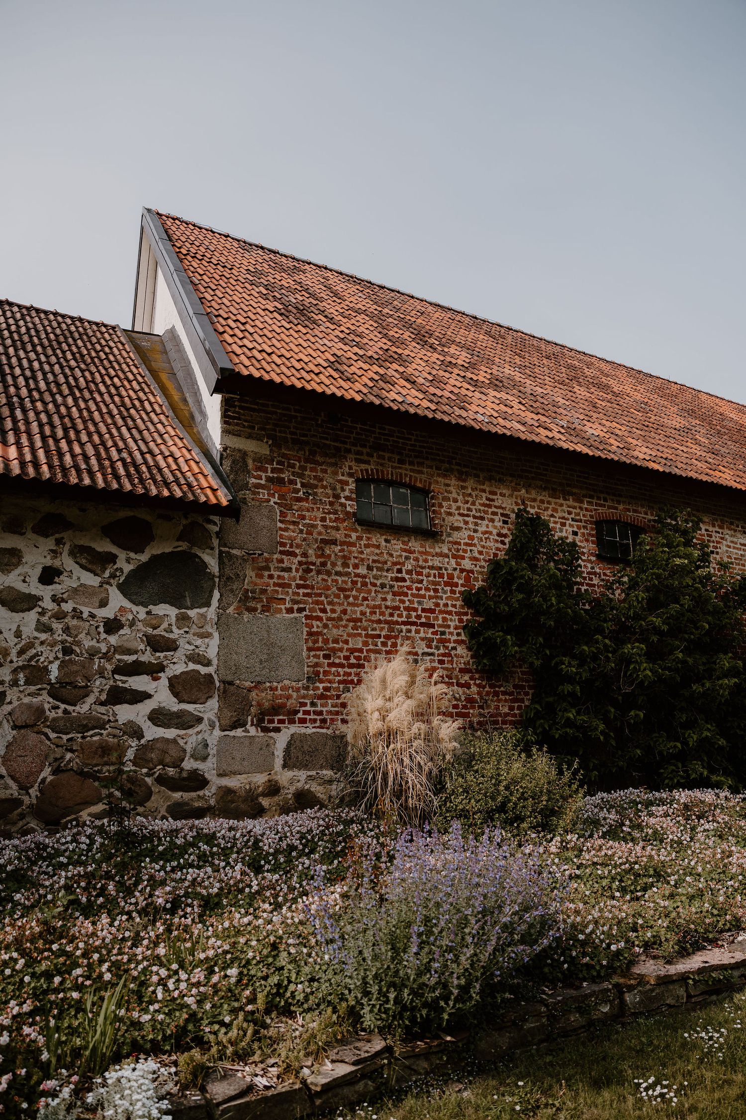 Rustic stone and brick barn with red tile roof and blooming wildflower garden against cloudy sky.