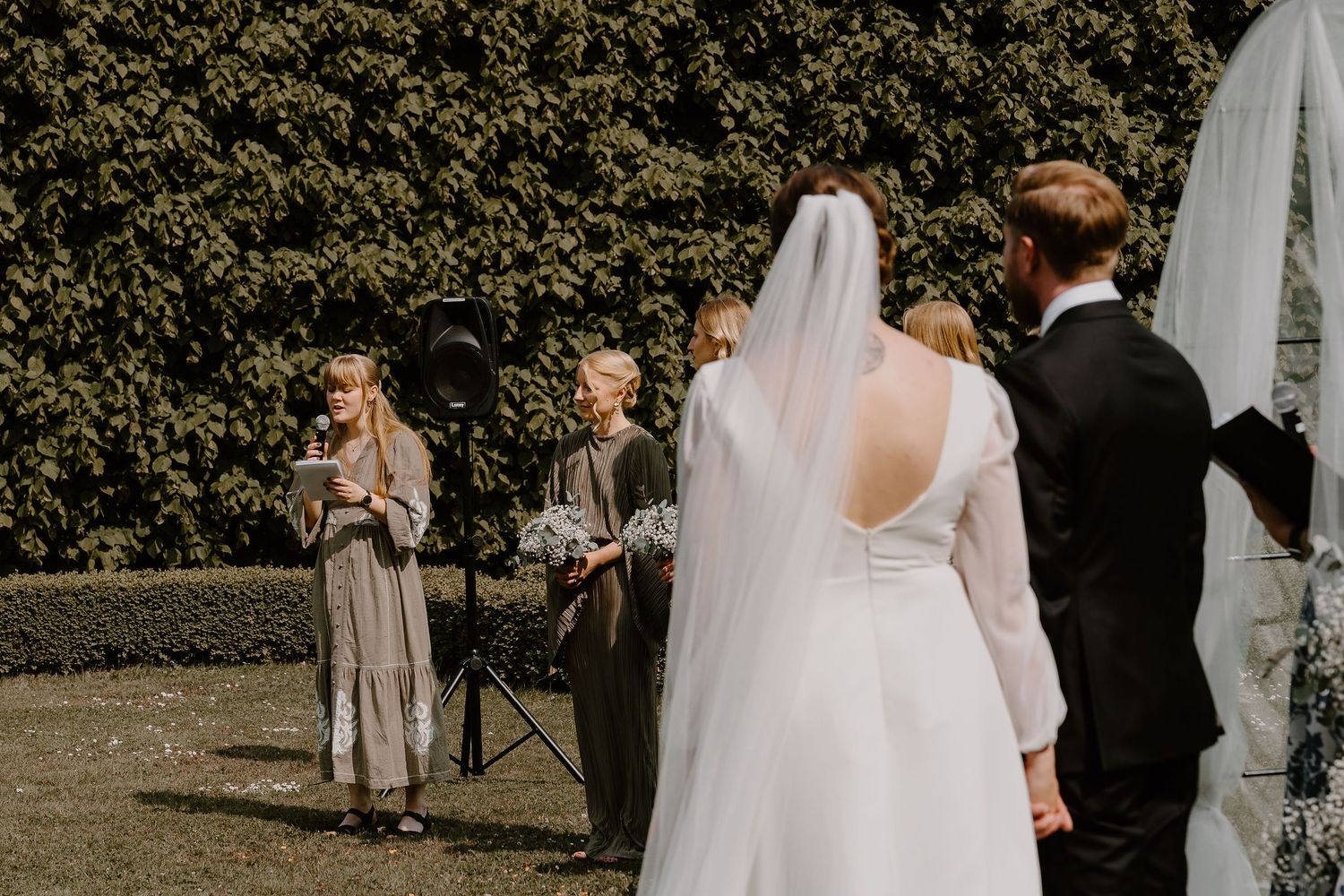 A wedding party gathers on a grassy lawn surrounded by trees and hedges on a sunny day.