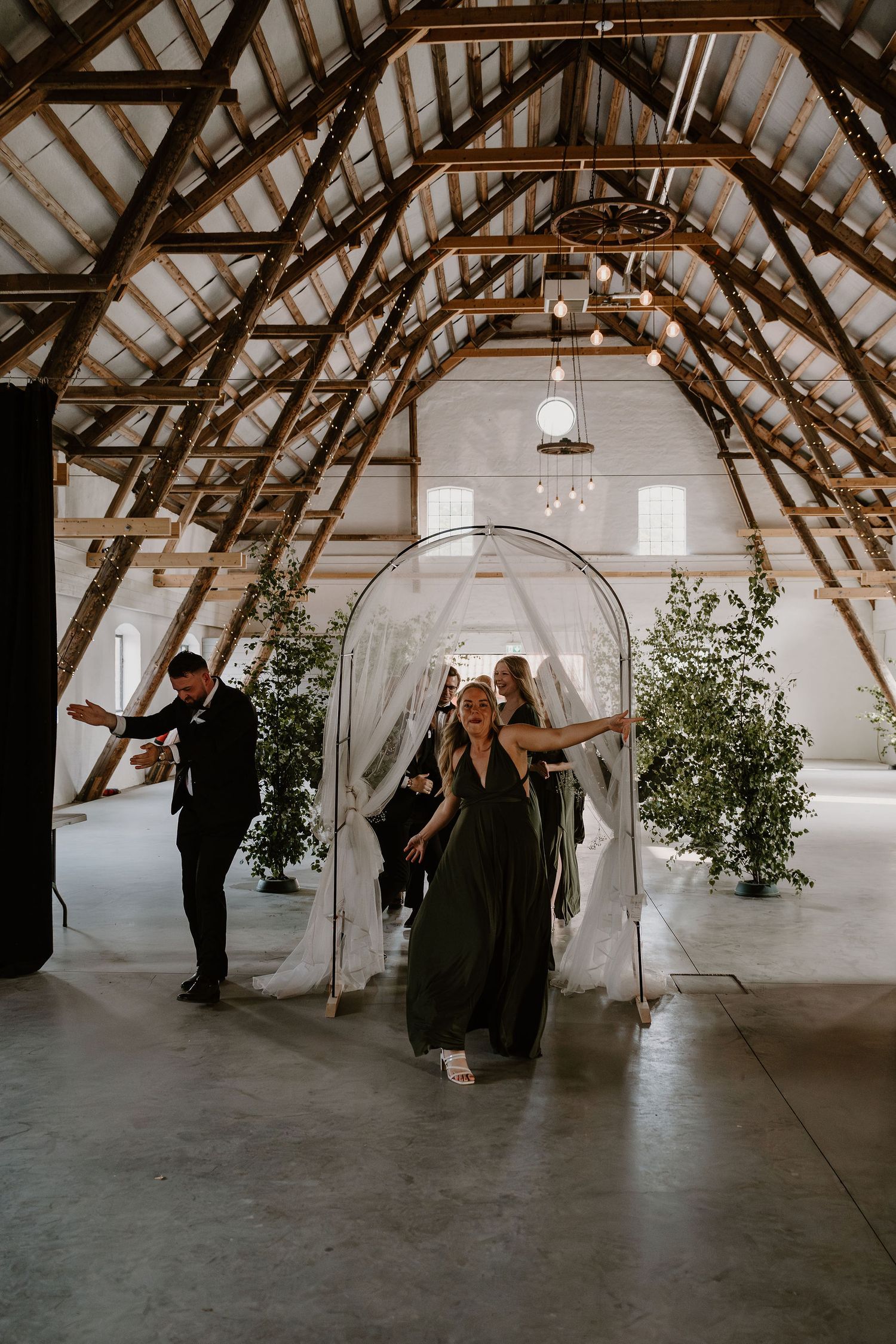 Wedding party in elegant black attire walking through a clear acrylic archway in a rustic barn venue with exposed beams.