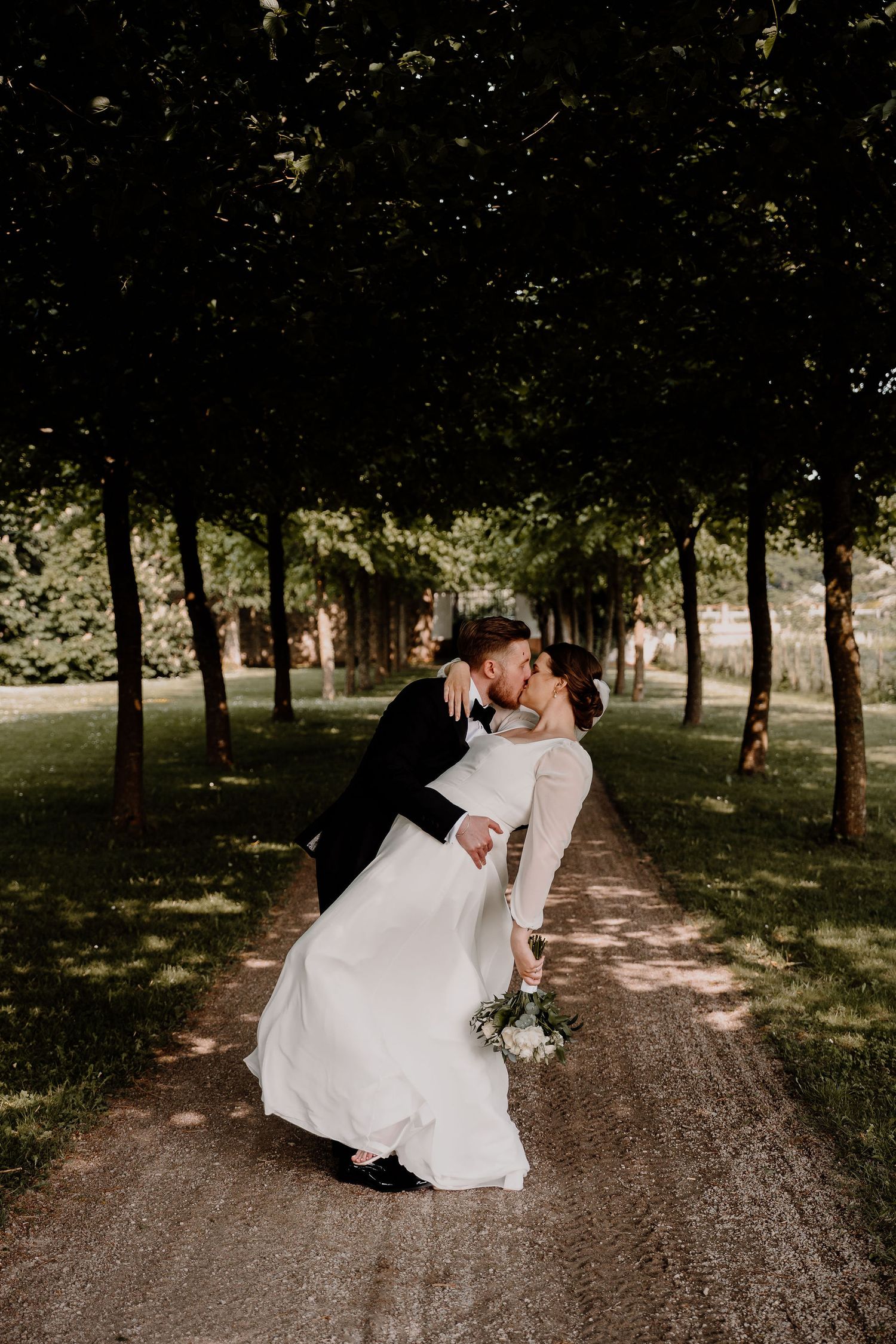 A romantic wedding scene with a couple sharing a kiss on a tree-lined path on their special day.