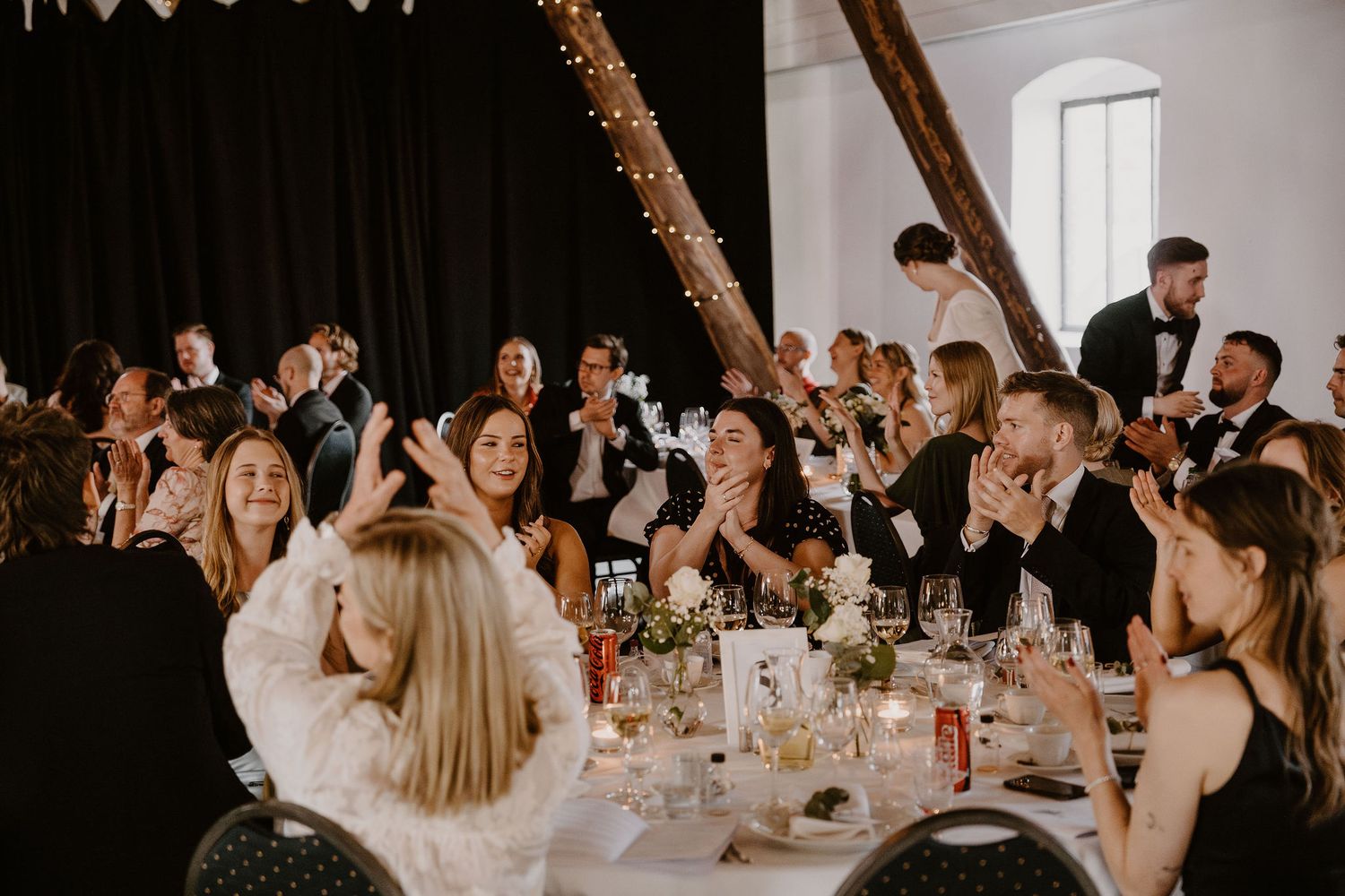 Wedding guests seated at long reception tables applaud during a speech in a rustic white barn venue.