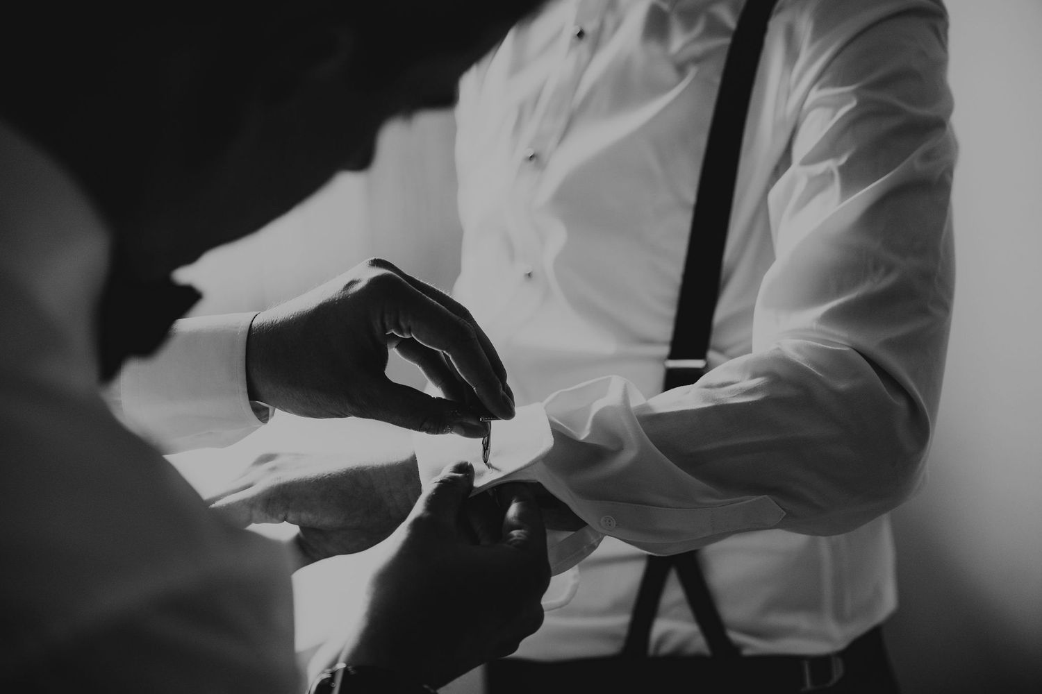 Black and white photo of hands wearing gloves receiving something from person in white dress shirt with suspenders.