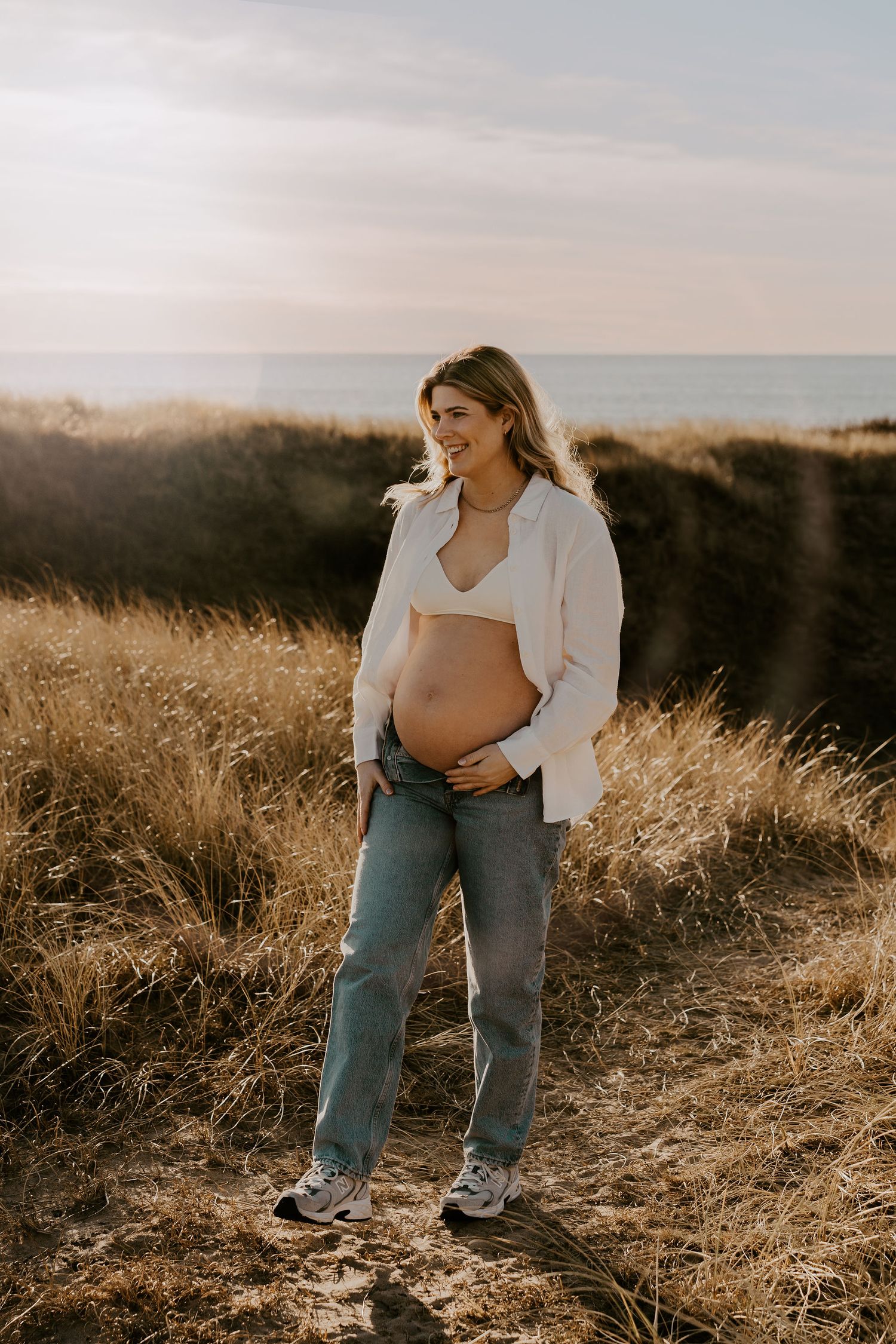 A pregnant woman stands on a hillside wearing jeans and an open white shirt at sunset with coastal waters in the distance.