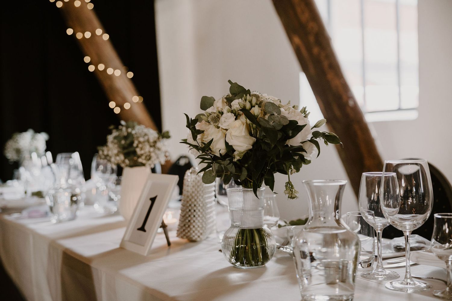 Elegant wedding table setting with white floral centerpieces, wine glasses, and string lights creating a romantic ambiance.