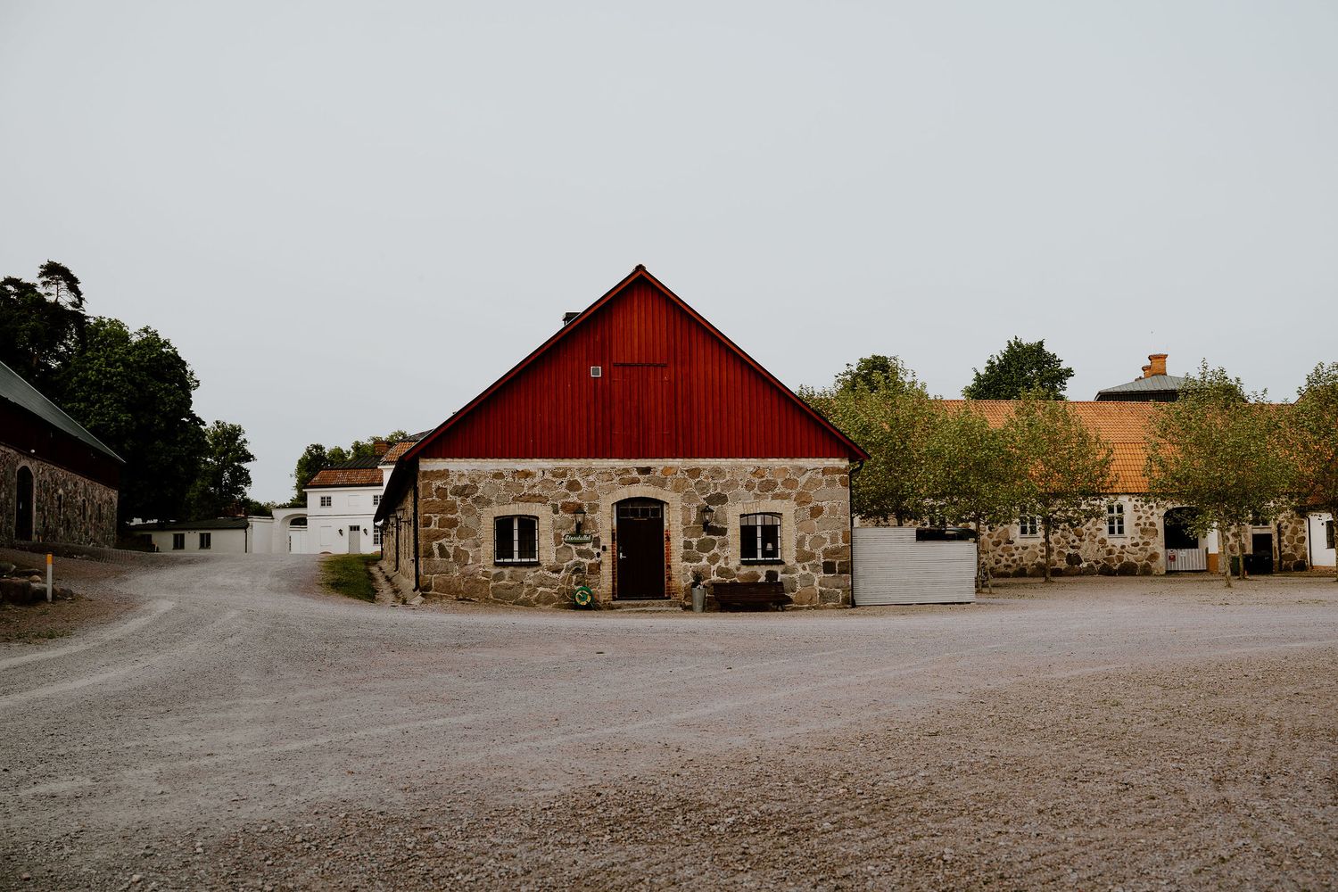 A rustic red barn with stone walls stands in a gravel courtyard surrounded by countryside buildings.