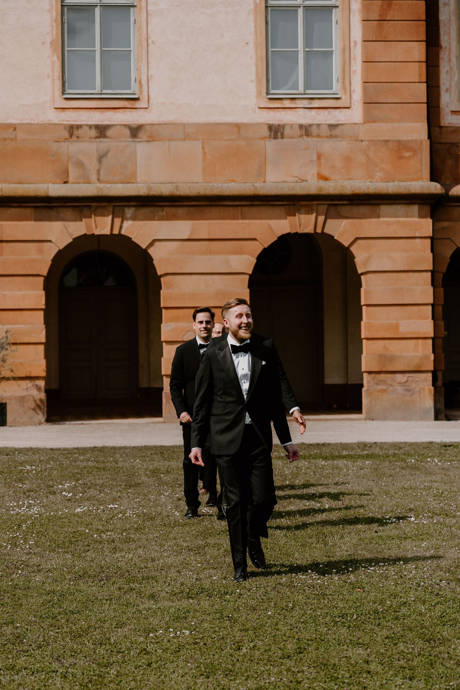 Two figures in formal black tuxedos walk on grass in front of a historic stone building with arched windows and pillars.