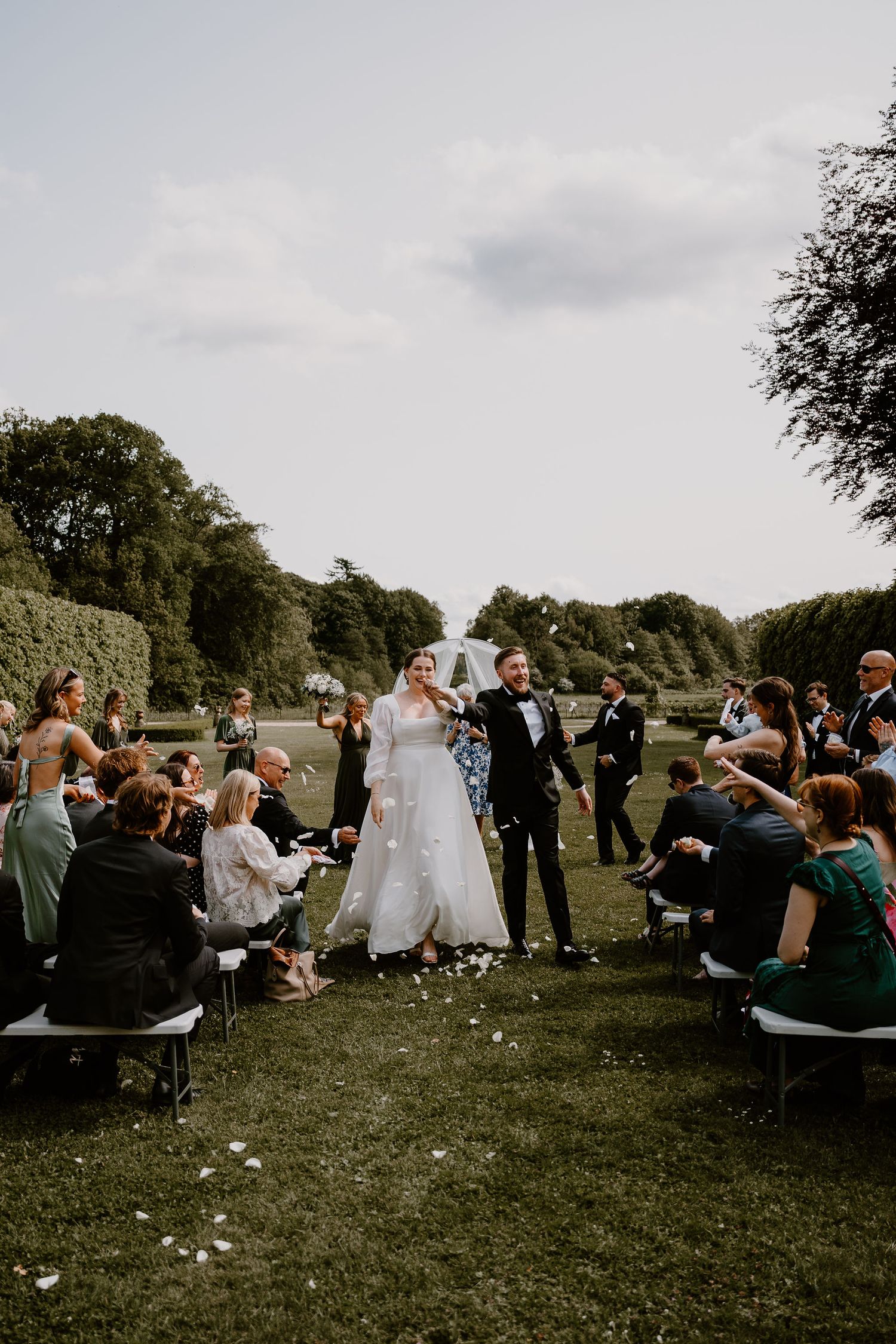 Wedding couple celebrates during outdoor ceremony with guests seated on benches in a garden setting while rose petals fall.