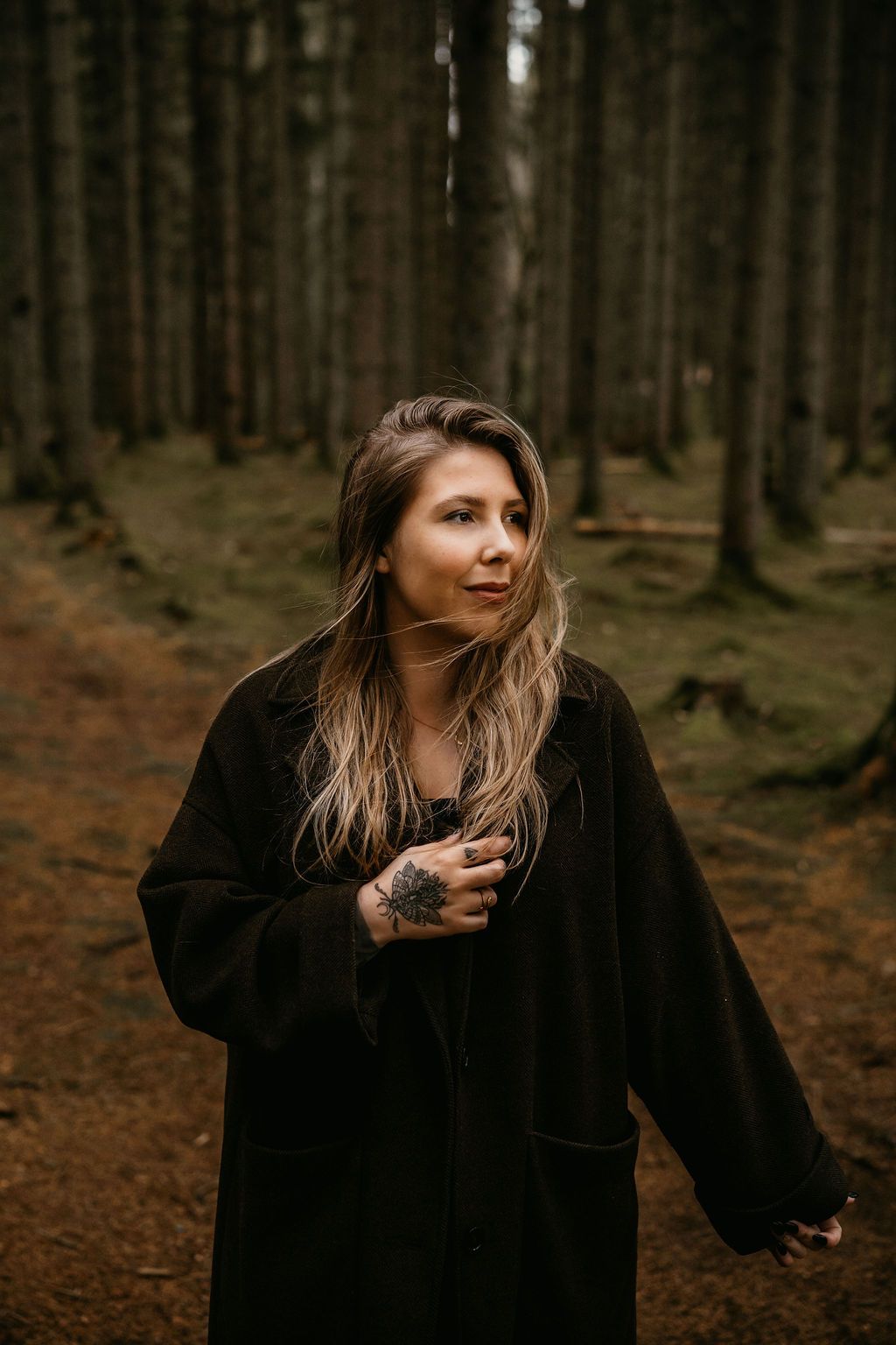 A person wearing a black coat stands amidst tall pine trees in a moody forest setting.