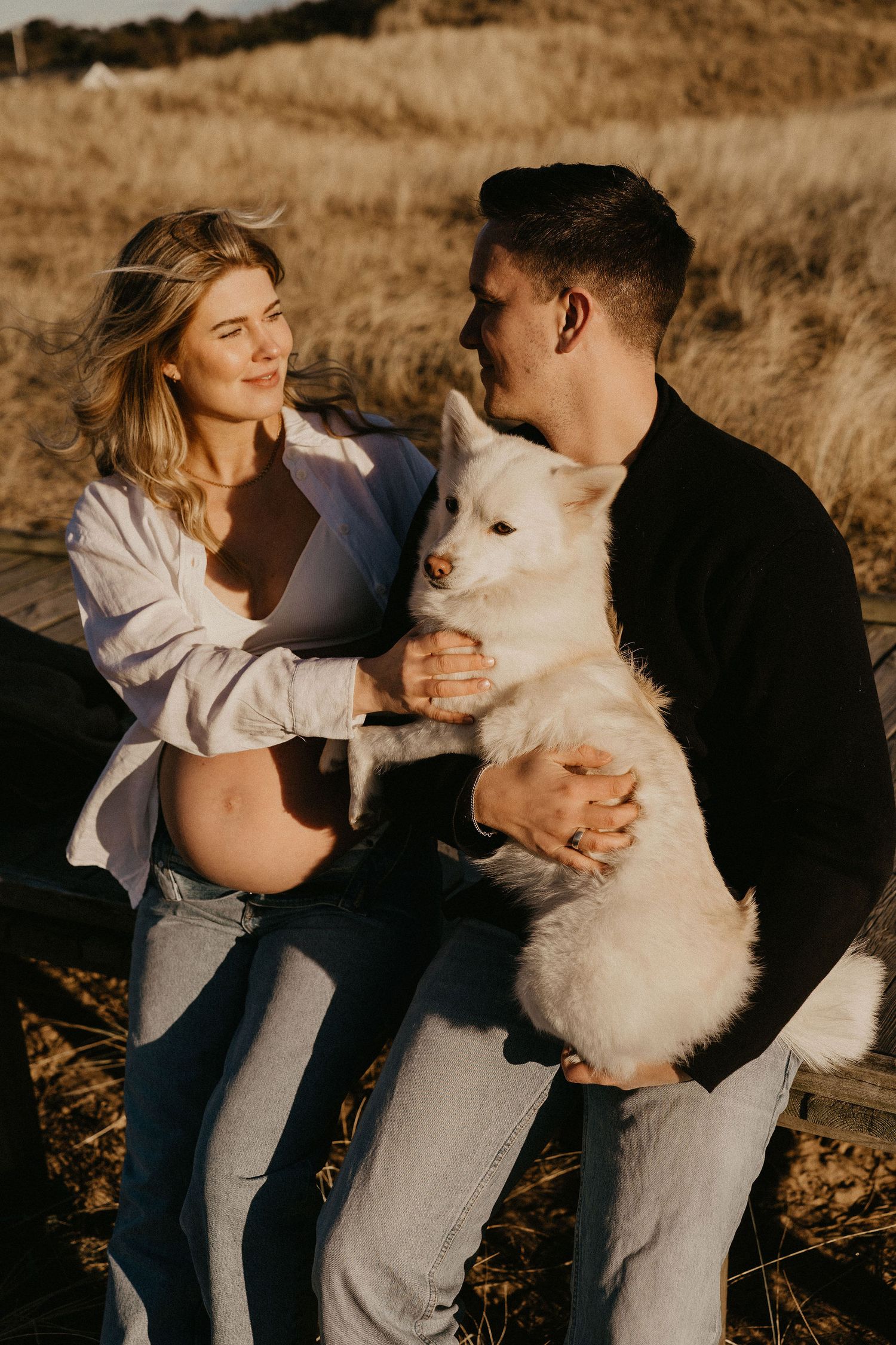 A couple sits outdoors at sunset with their fluffy white dog, sharing an intimate moment in nature.