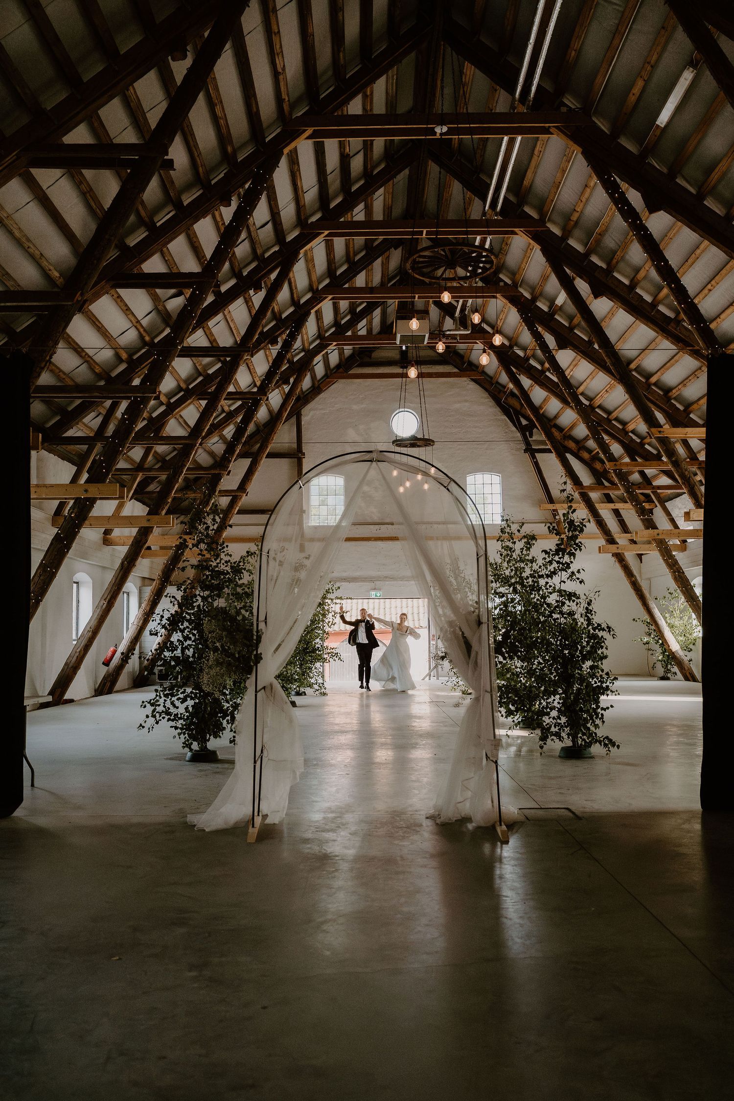 Elegant barn venue interior with exposed wooden beams and white fabric arch decorated with greenery for a wedding ceremony.