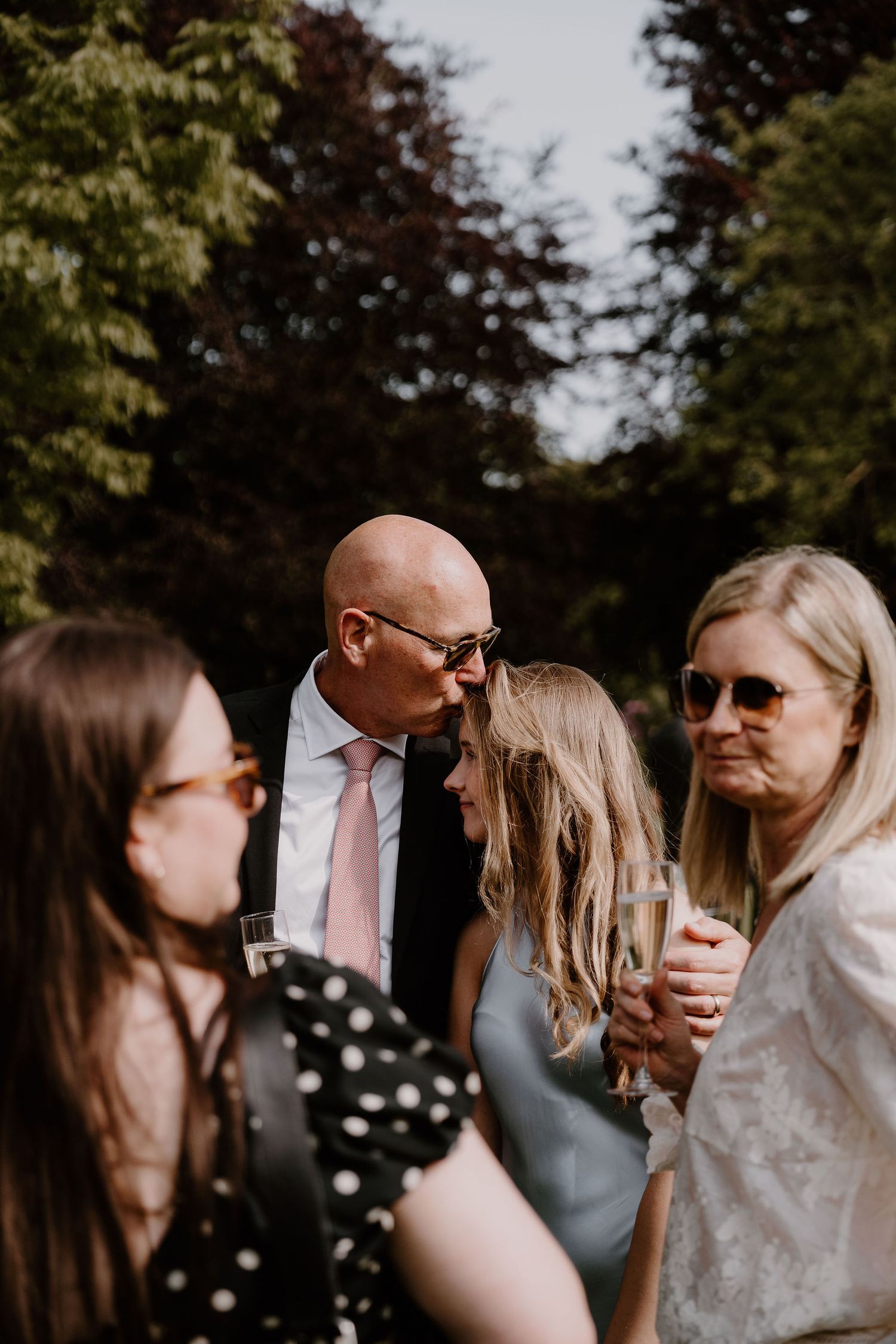 A group of people wearing sunglasses gather together outdoors with lush green trees in the background.
