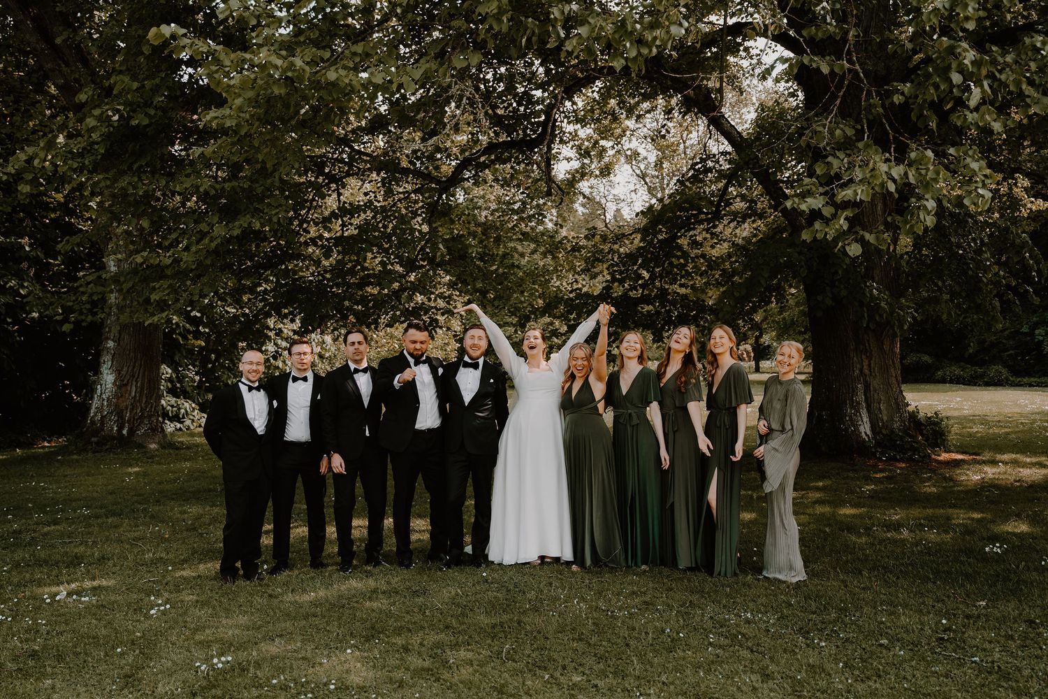 Wedding party in formal attire poses together outdoors under tall trees on a sunny day.