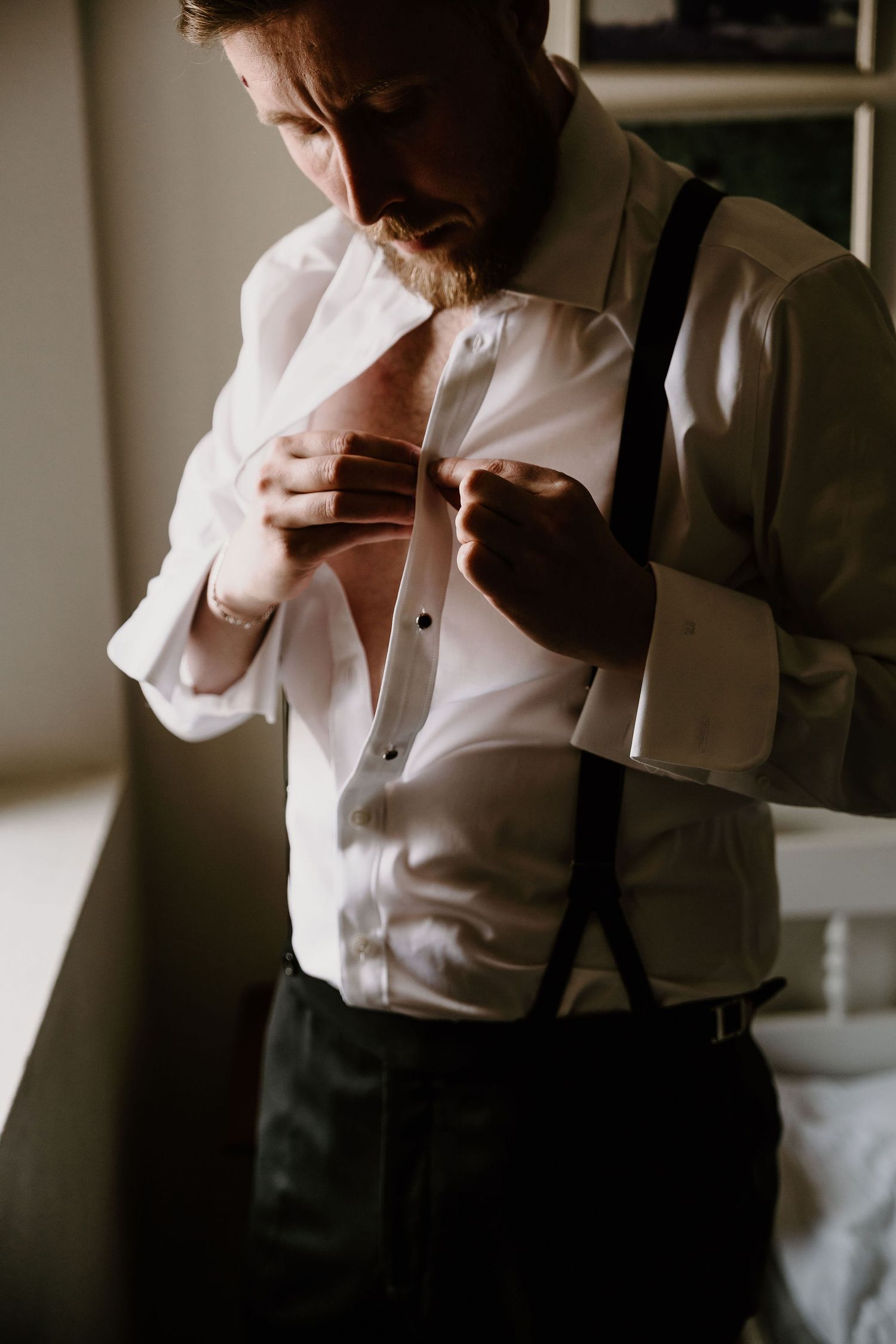 A person in formal attire adjusts their suspenders while standing in a dimly lit room.