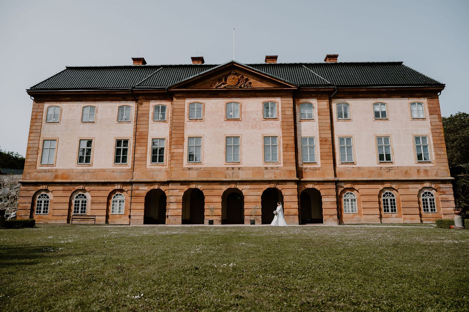 A grand historic manor building with pale orange facade and black roof features arched colonnade and symmetrical windows.