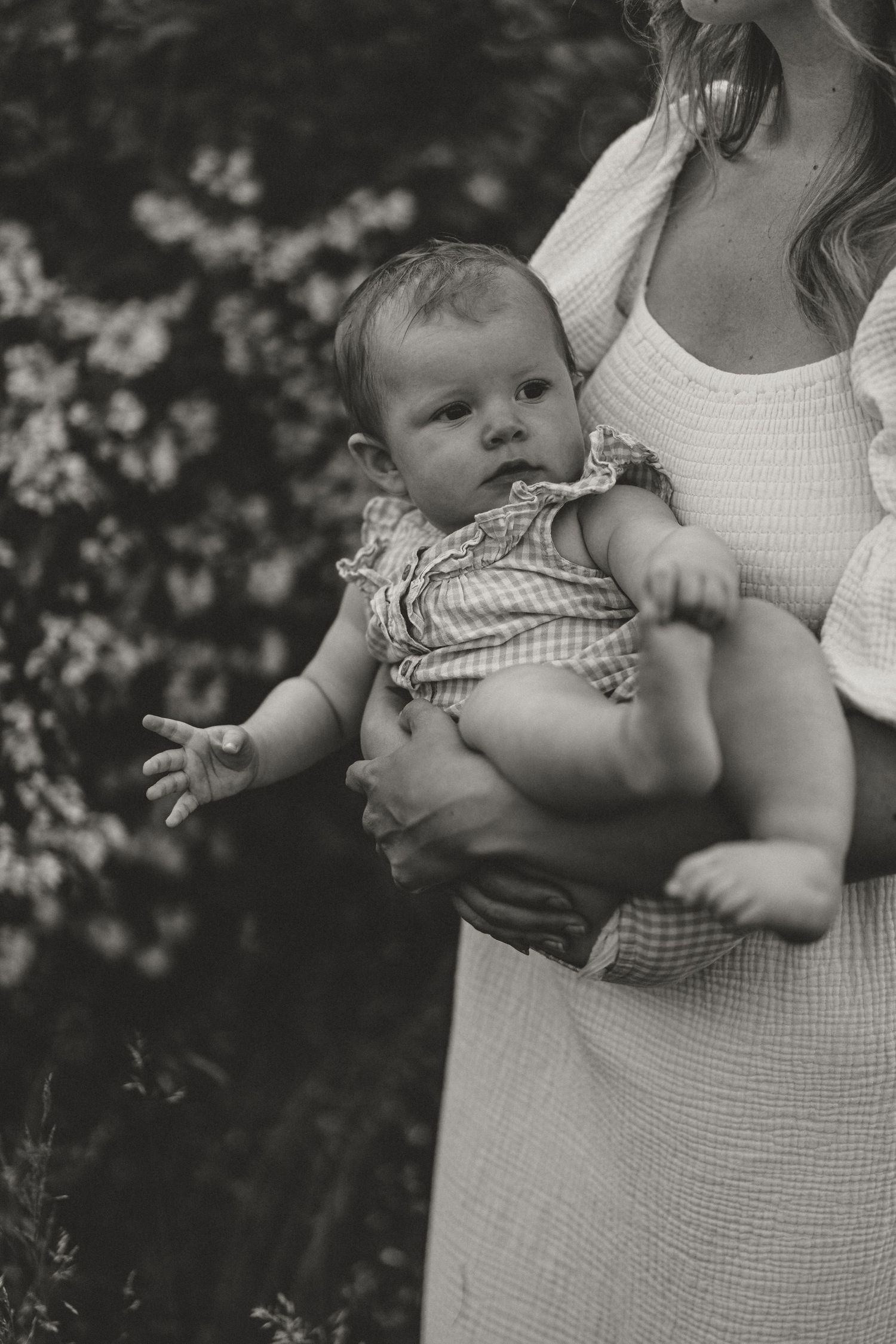 Black and white photo of a baby in a gingham romper being held by someone wearing a white dress in a garden setting.
