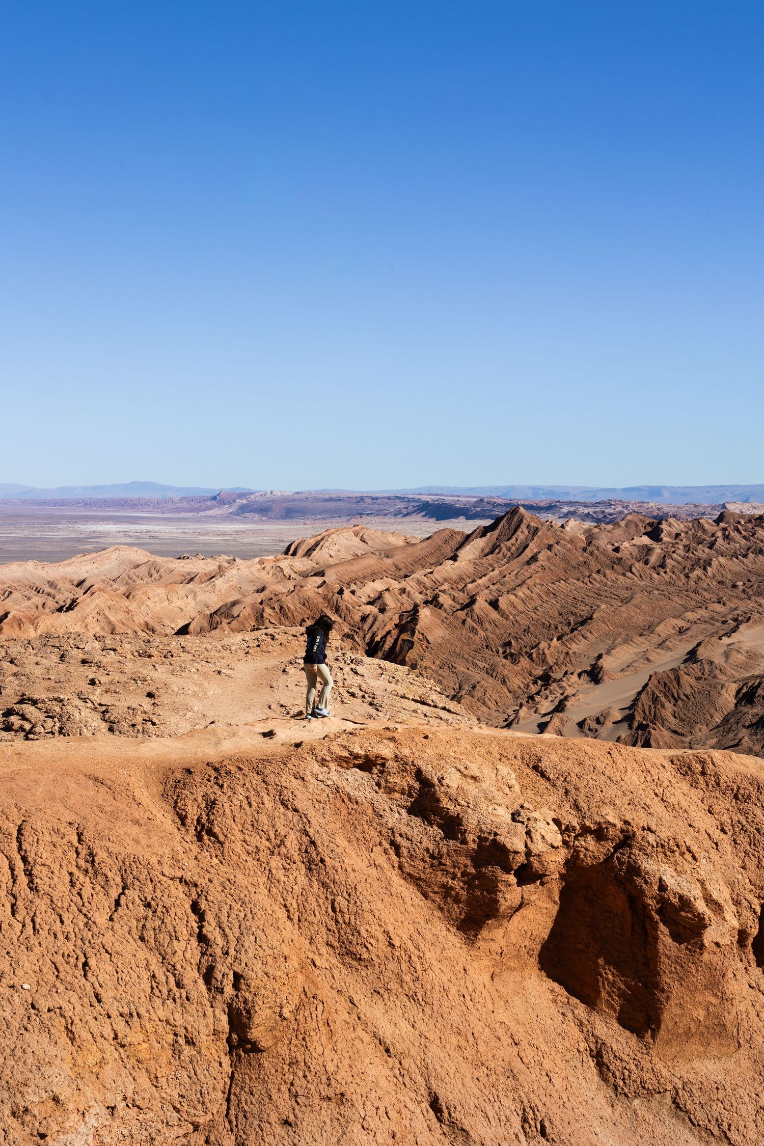 A lone hiker stands at the edge of a vast desert canyon overlooking the rocky terrain and blue sky in Valle de la Luna, Chile.