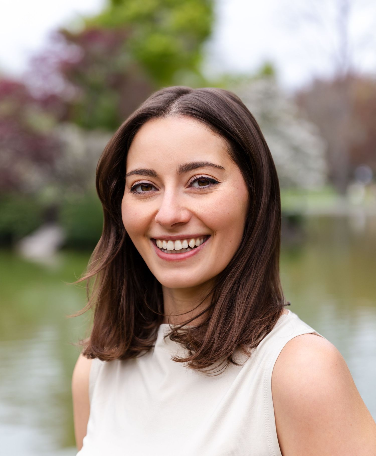 Person in white sleeveless top smiles outdoors with water and trees in background.