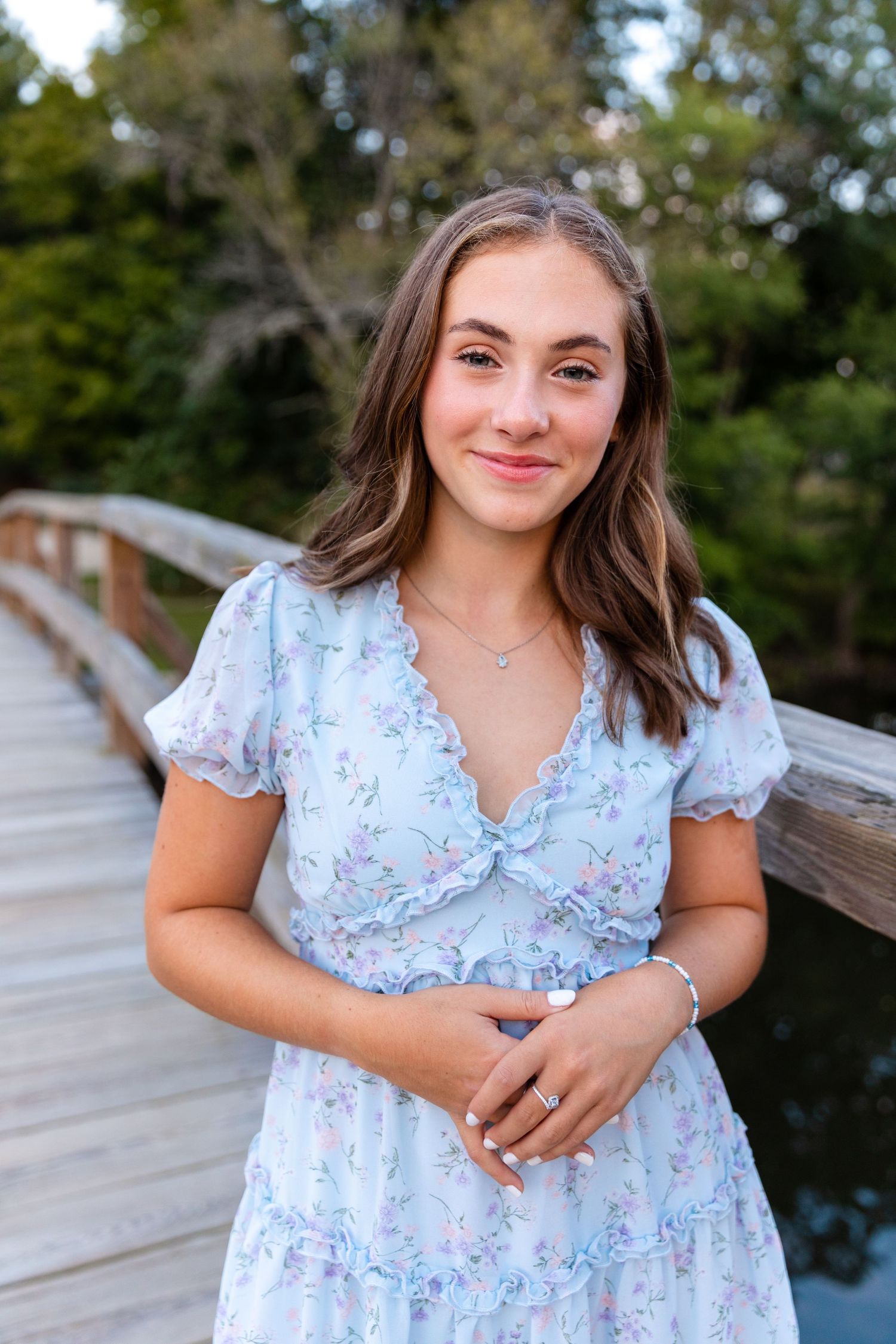 Portrait of young woman in floral dress standing on wooden boardwalk near water.